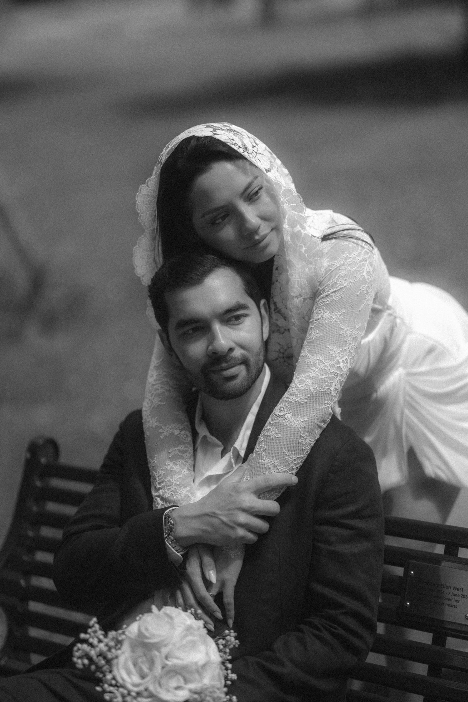 Black and white wedding photo of bride in lace veil embracing groom on bench, holding white rose bouquet.