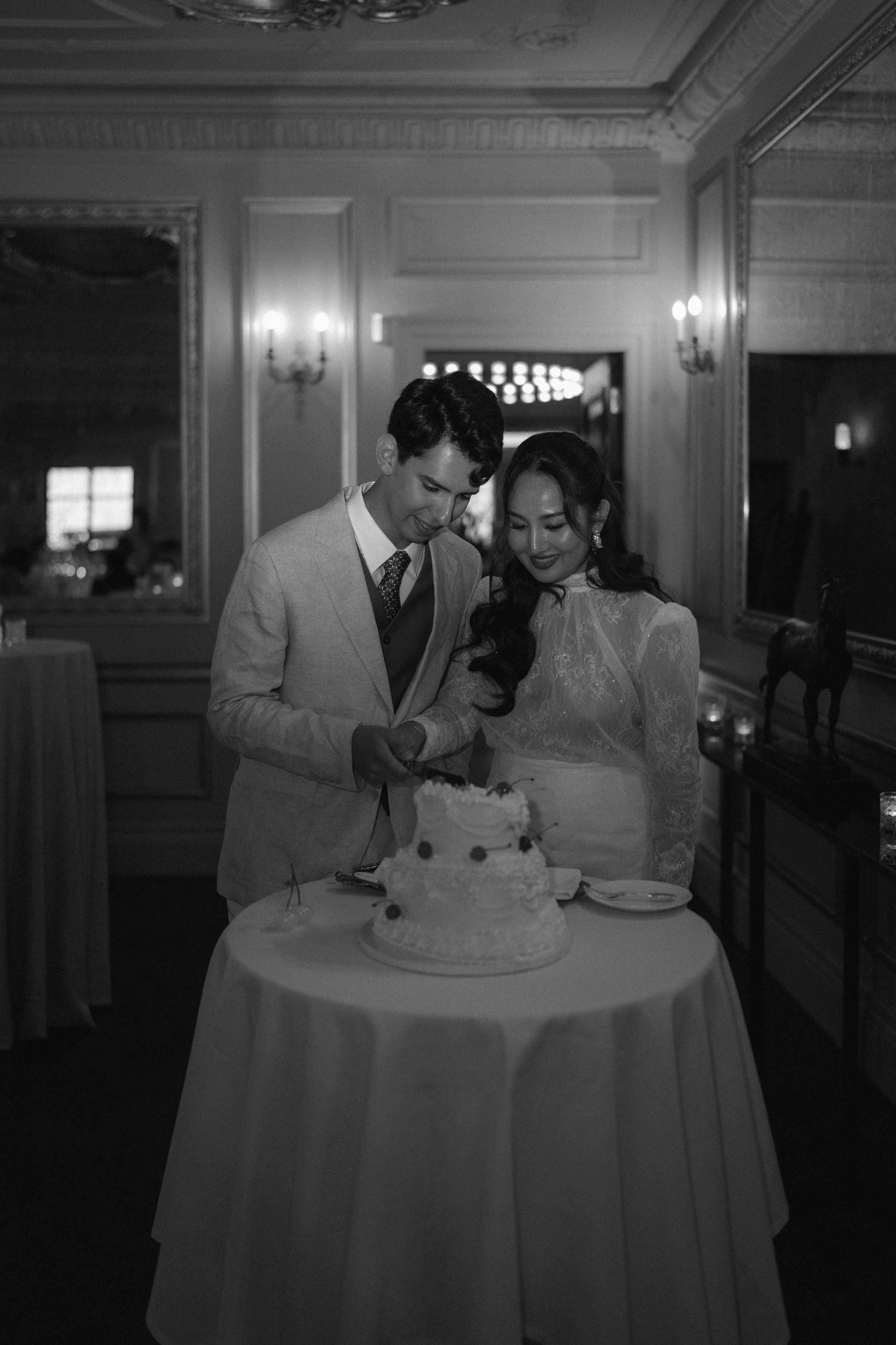 A couple in formal attire cuts their wedding cake together at an elegant reception venue in black and white photography.