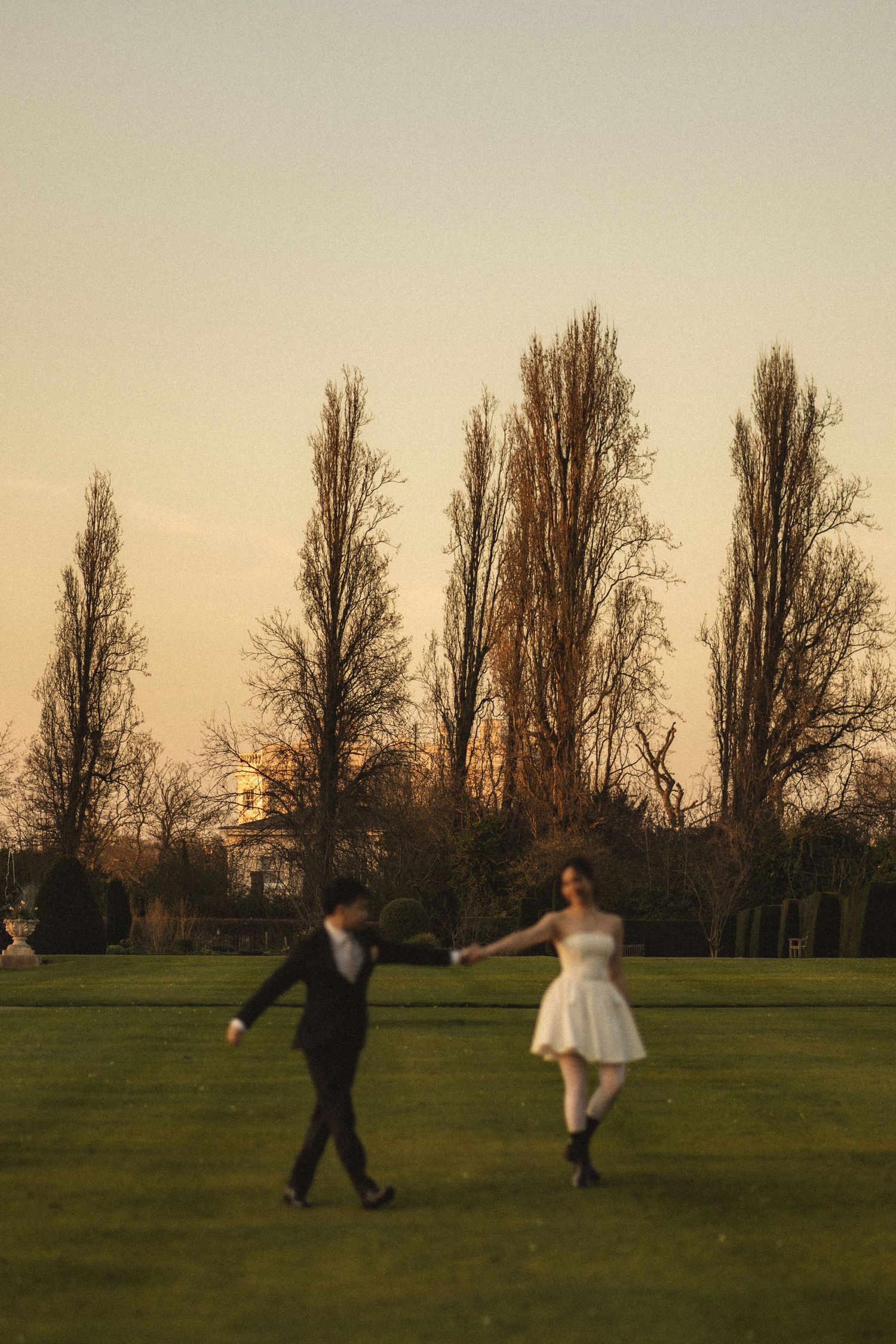 Two people run joyfully across a grassy field at sunset with tall trees silhouetted in the background.