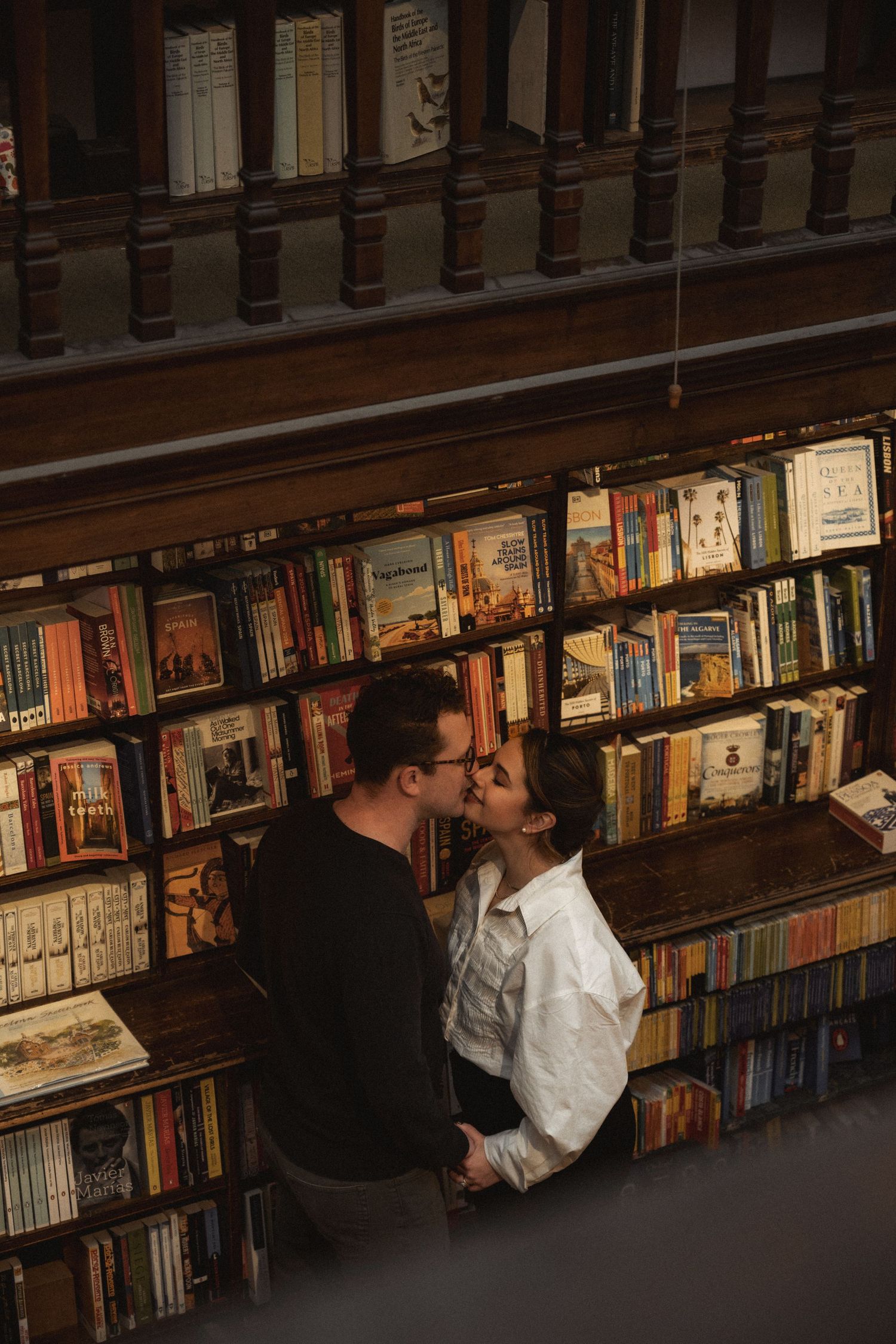 A couple shares an intimate moment between bookshelves in a dimly lit library or bookstore.