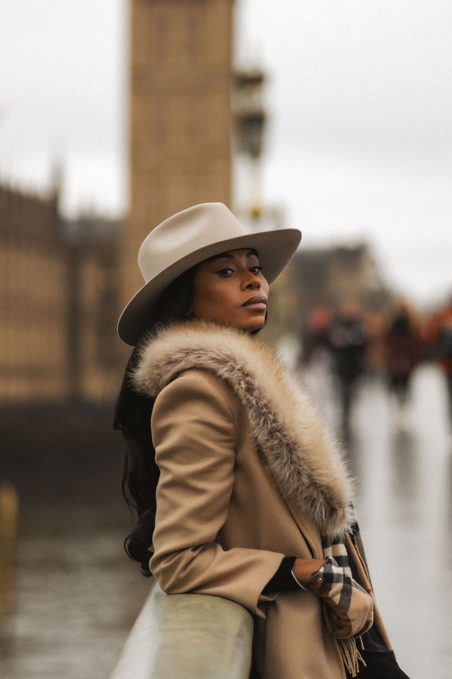 A person in a beige coat with fur trim and wide-brimmed hat poses against a blurred urban backdrop.