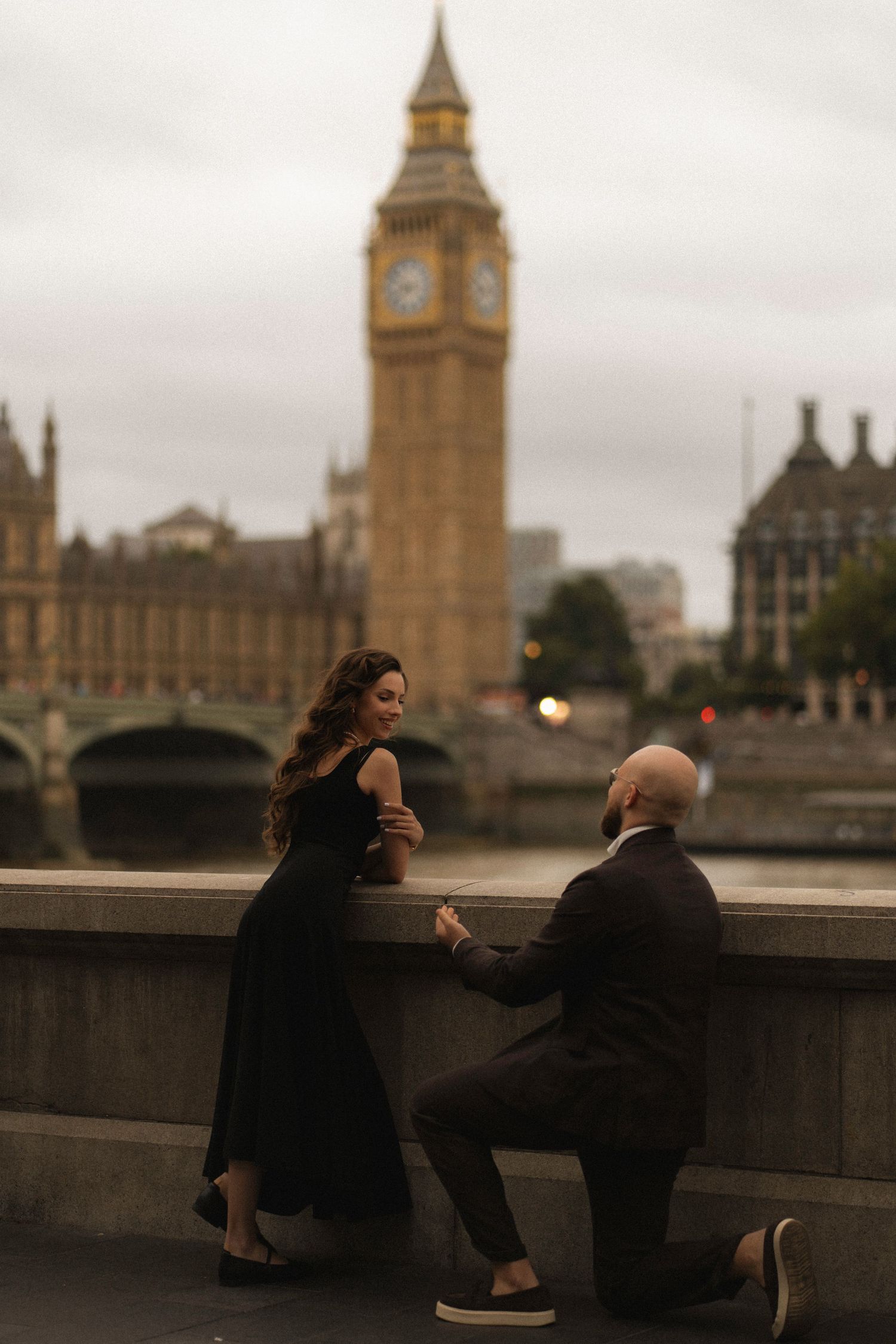 Man proposing to woman in black dress with Big Ben and Westminster Bridge in background at dusk in London.