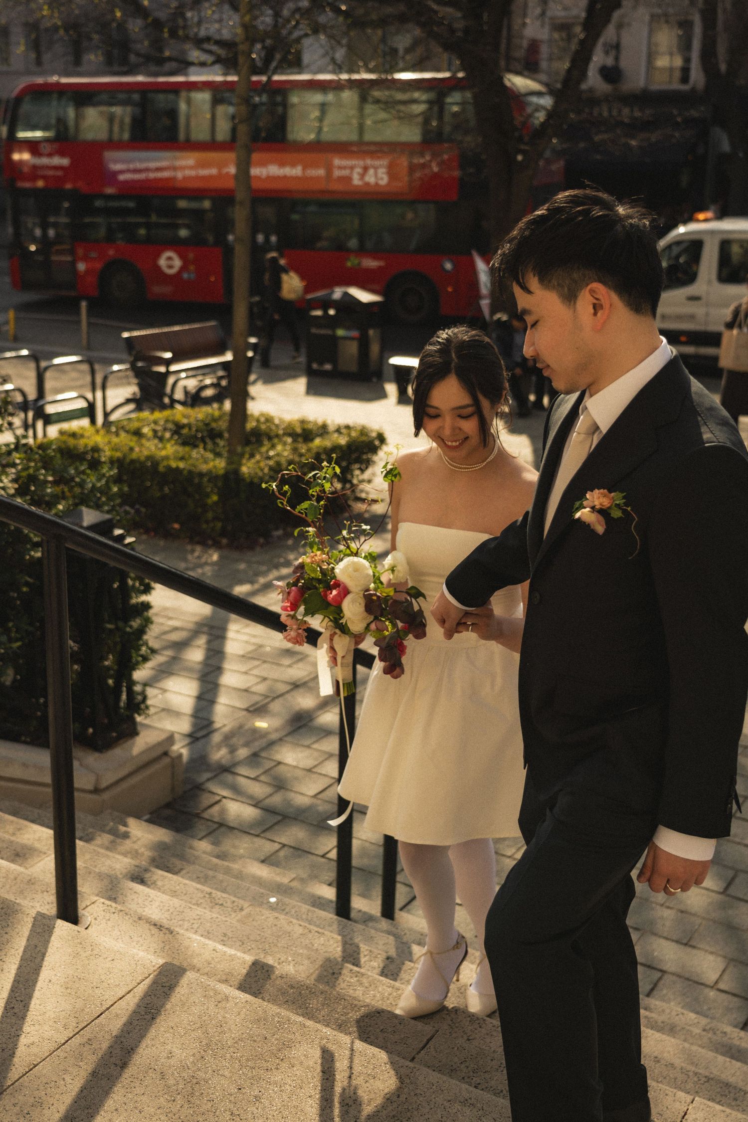 A couple in wedding attire walks down outdoor steps in an urban setting while holding a floral bouquet.