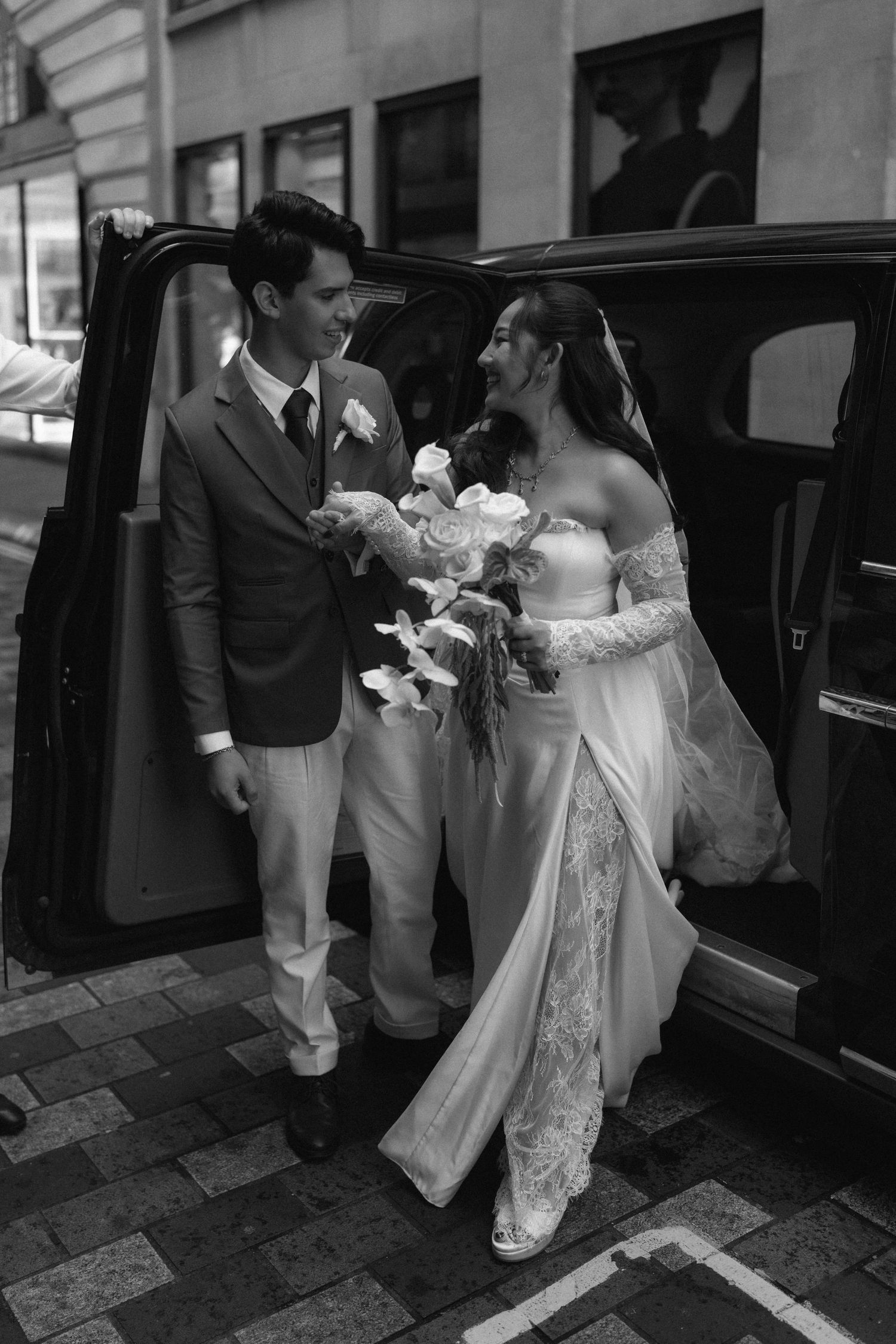 A bride in a white lace dress is being helped into a black car by wedding attendants in formal attire.