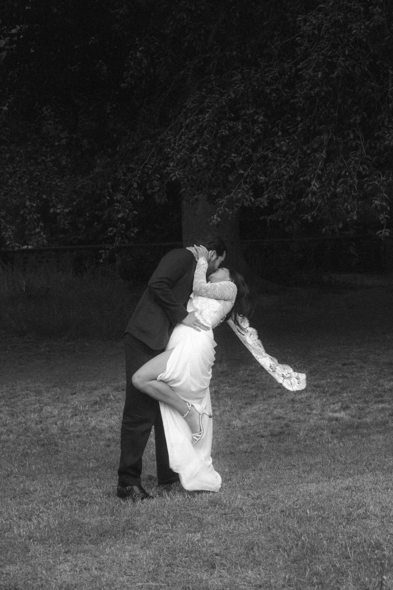 Black and white photo of a bride and groom sharing a romantic dip kiss on a grass lawn with trees in the background.