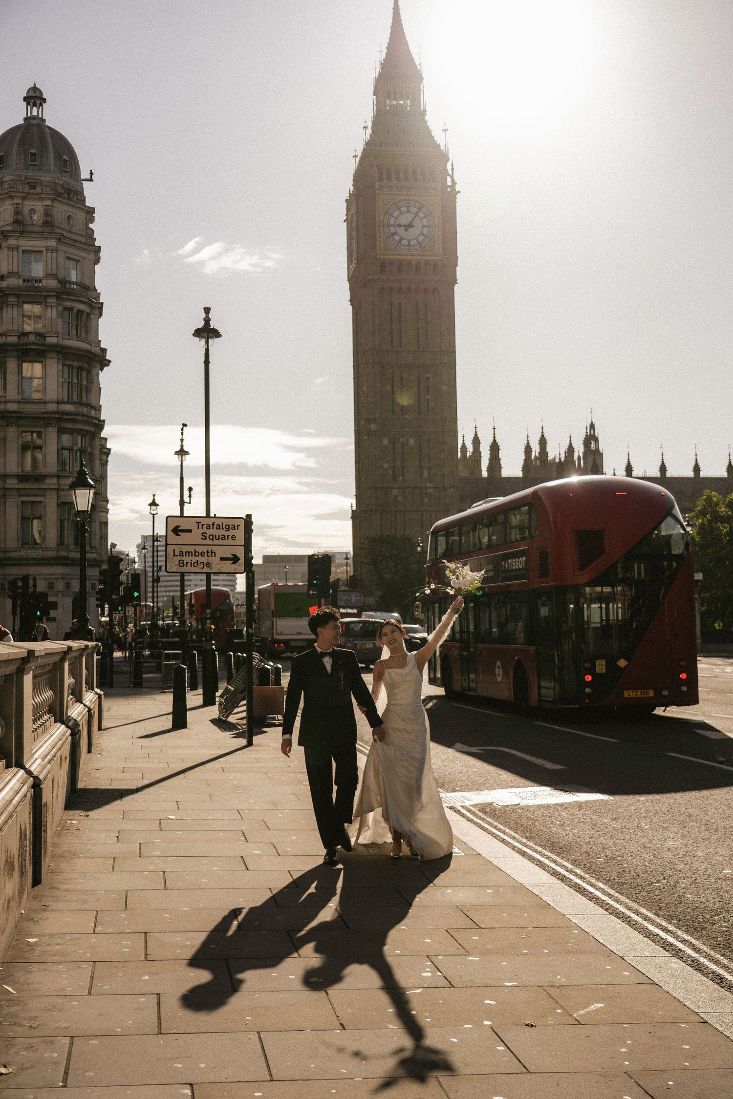 Bride and groom walk near Big Ben with a red double-decker bus in London on their wedding day.