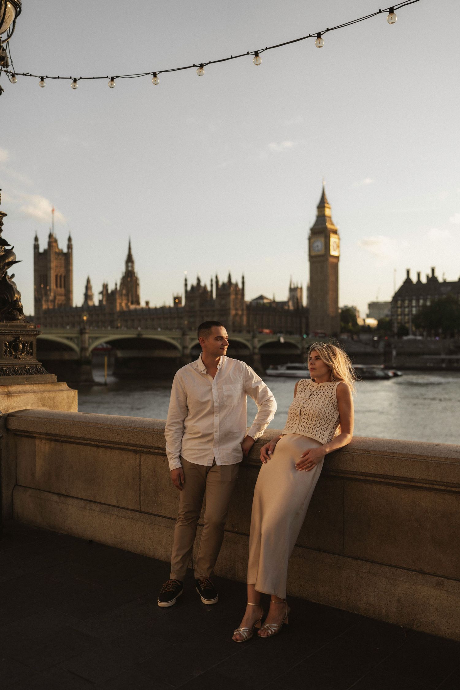 A couple sits on a stone wall at sunset with London's iconic Houses of Parliament and Big Ben silhouetted in the background.