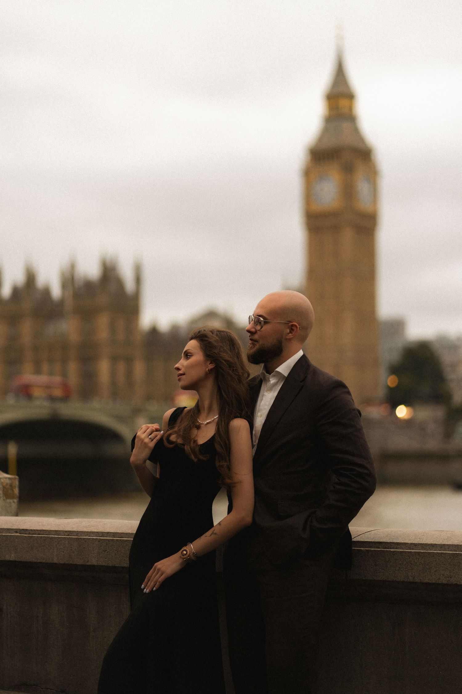 A couple in formal attire poses romantically by the Thames River with Big Ben and Westminster in the background at dusk.