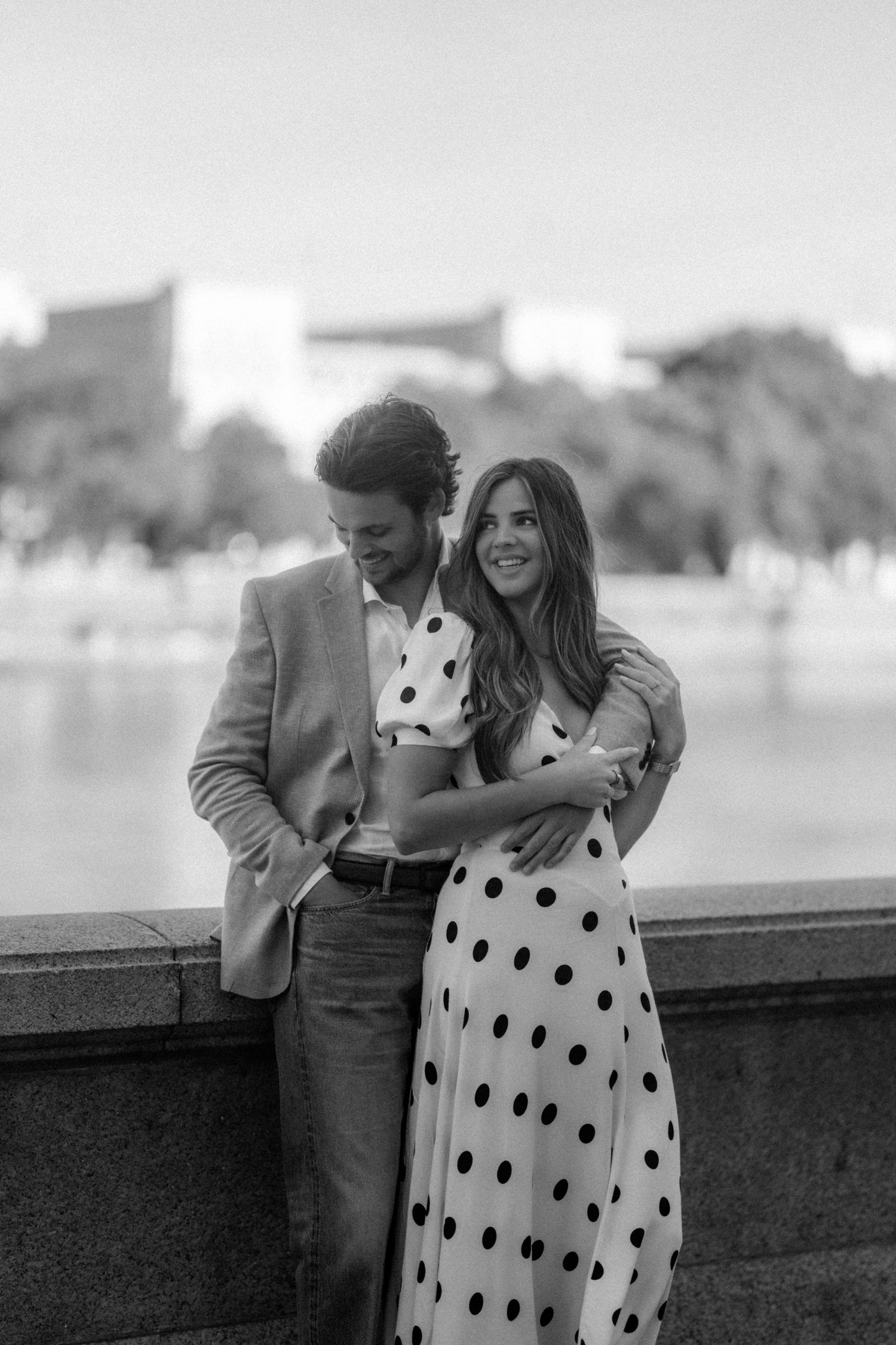 Black and white photo of a romantic couple embracing outdoors, woman in polka dot dress, man in blazer, near a waterfront.