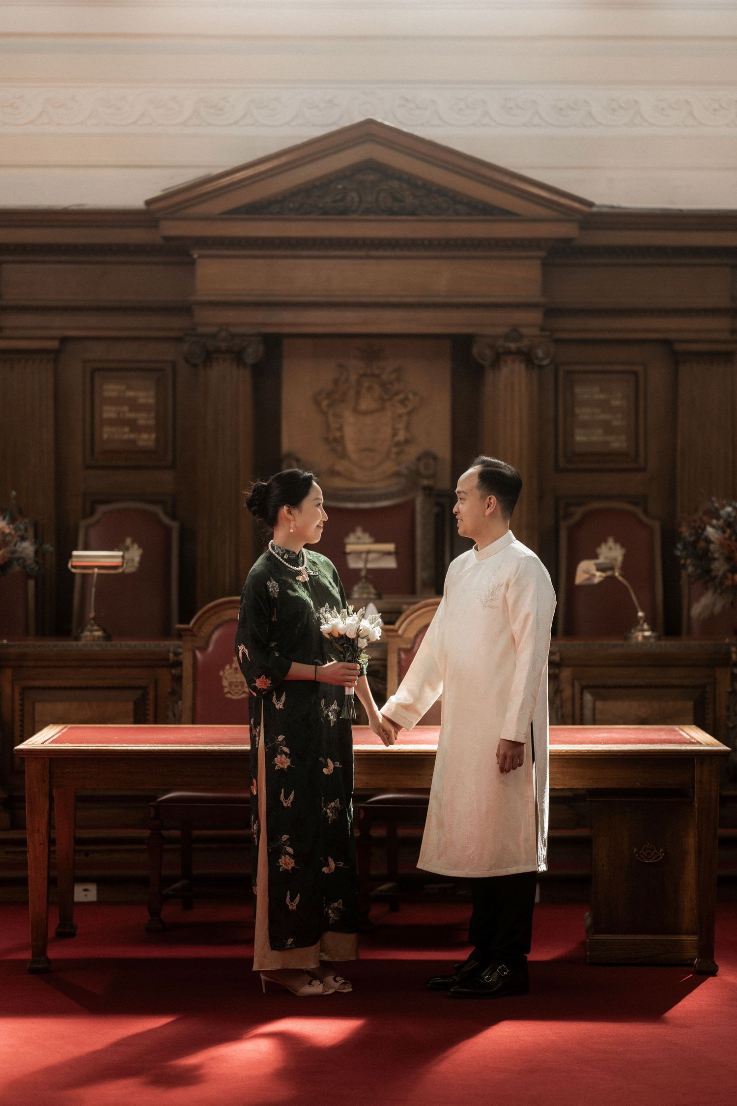 Couple in traditional Vietnamese ao dai attire holding hands in an elegant wood-paneled ceremonial hall.
