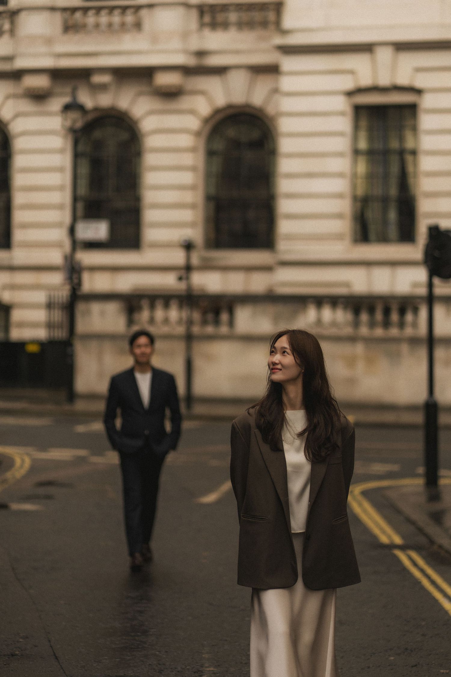 A person in a brown blazer and white outfit stands on a city street with classical architecture in the background.