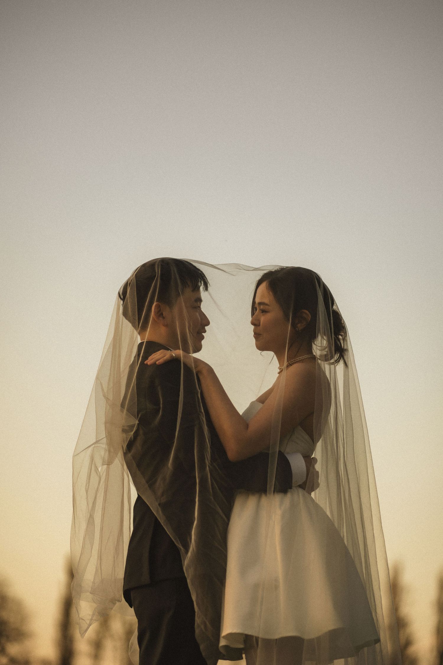 A couple shares an intimate moment under a bridal veil at sunset, the bride in white and groom in a dark suit.