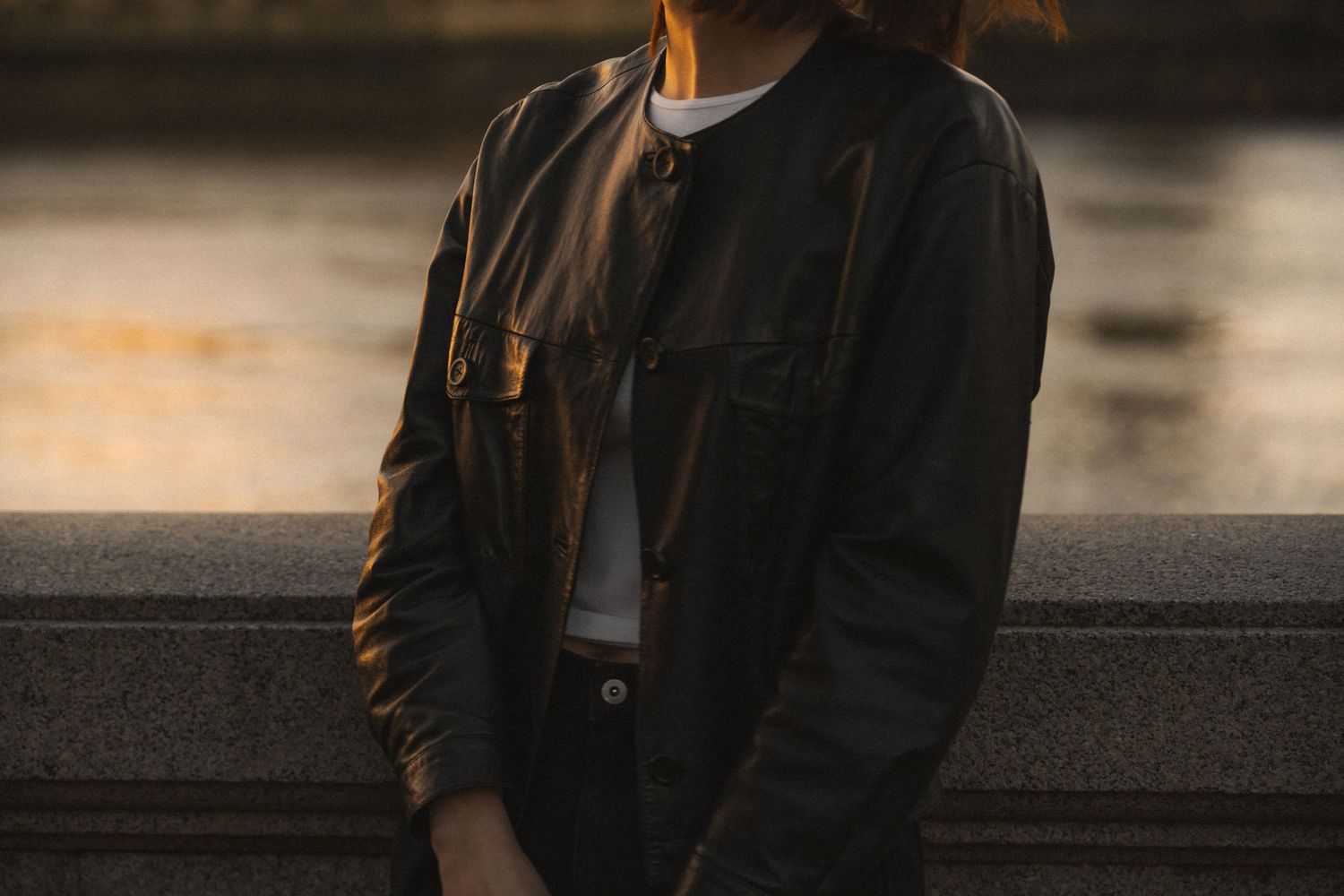 Person in dark jacket stands against blurred urban background at sunset with warm lighting.