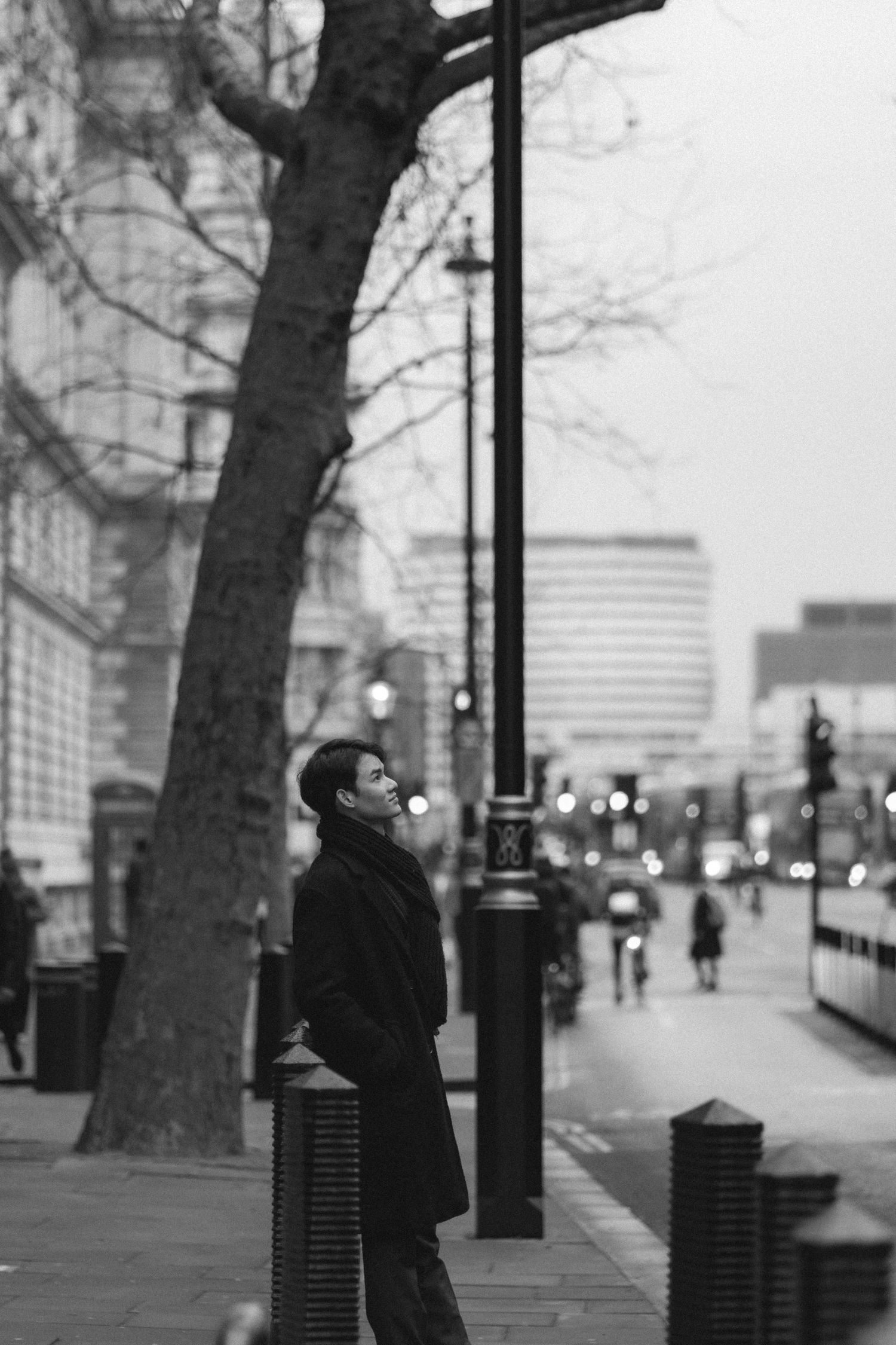 A person in dark clothing gazes upward while standing on an urban street with trees and buildings in the background.