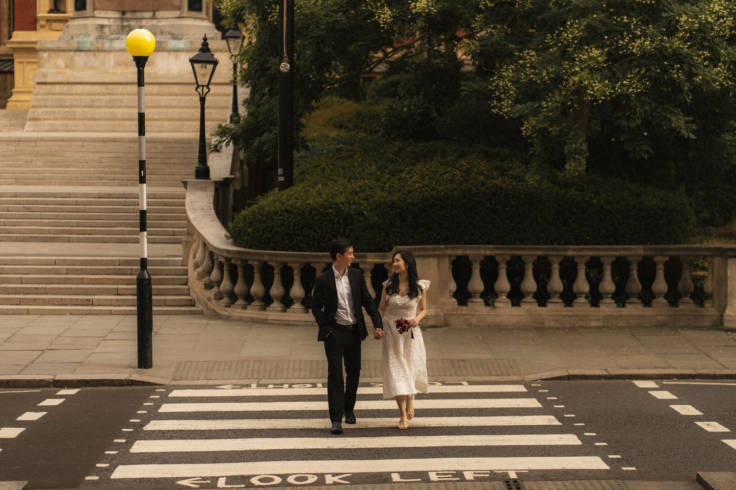A couple walks hand in hand across a crosswalk near an elegant stone staircase with ornate balustrades and lamp post.