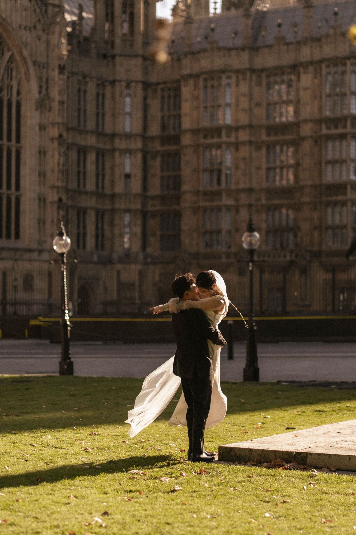 A couple embraces in wedding attire on the lawn outside the Houses of Parliament in London on a sunny day.