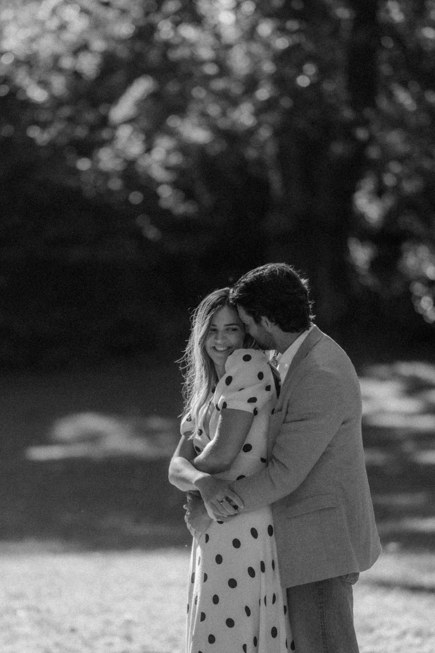 Black and white photo of a smiling couple embracing outdoors, woman in polka dot dress, man in suit jacket.
