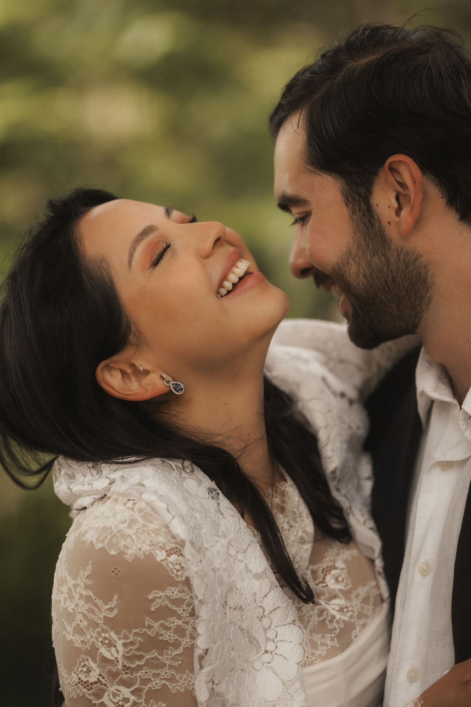 A joyful couple in wedding attire laughing together outdoors, bride in lace dress with dangling earrings.
