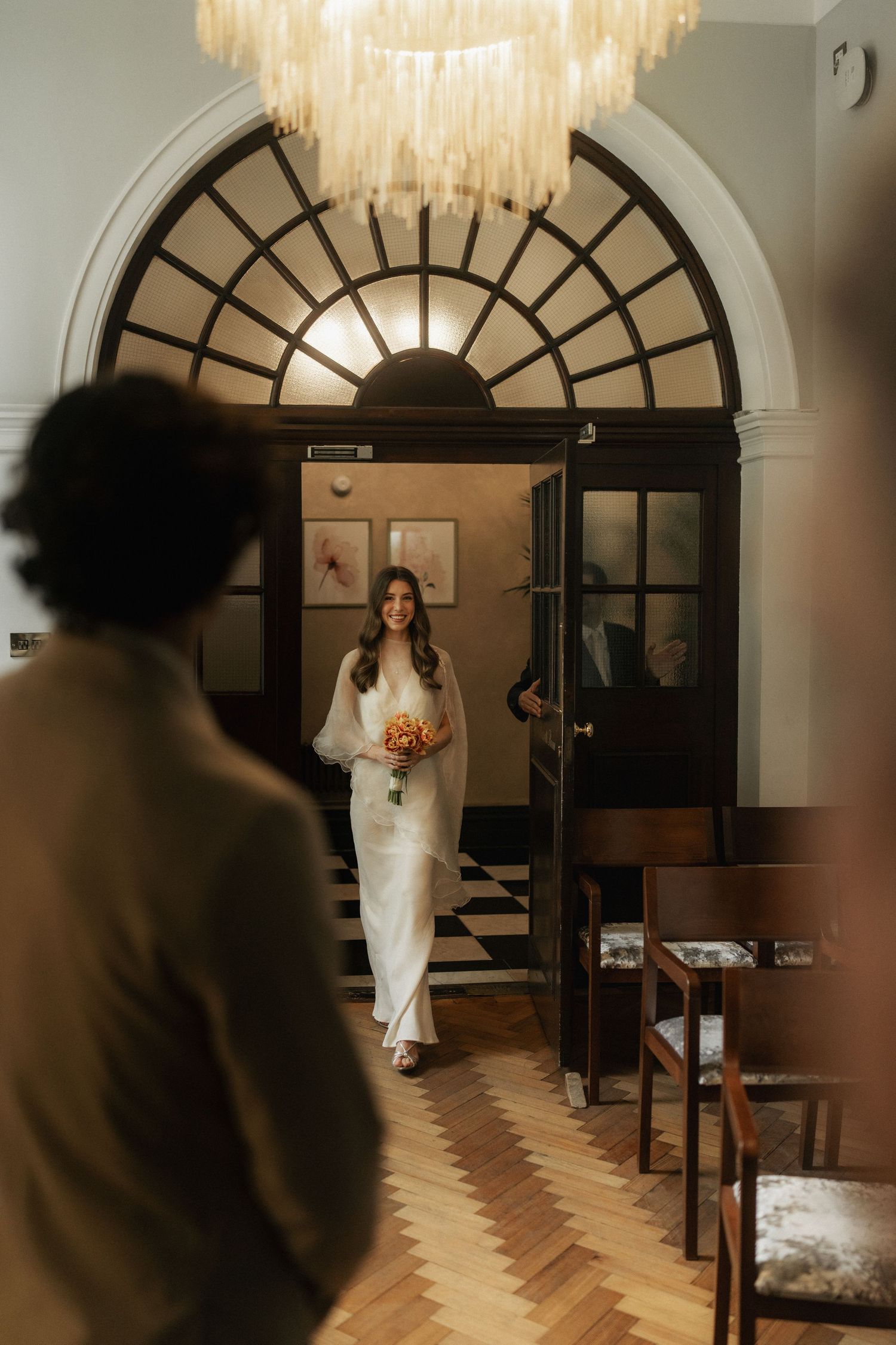 Bride in white gown holding bouquet walks through arched doorway toward groom in elegant venue with chandelier.