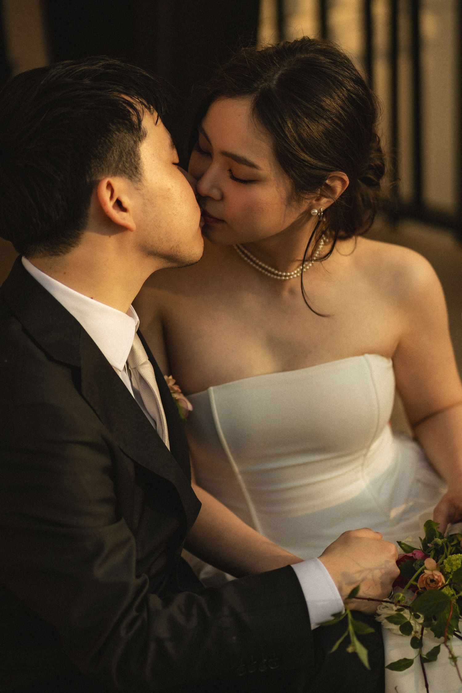 A bride and groom share a romantic kiss in warm golden light, she wearing a strapless white gown and pearl necklace.