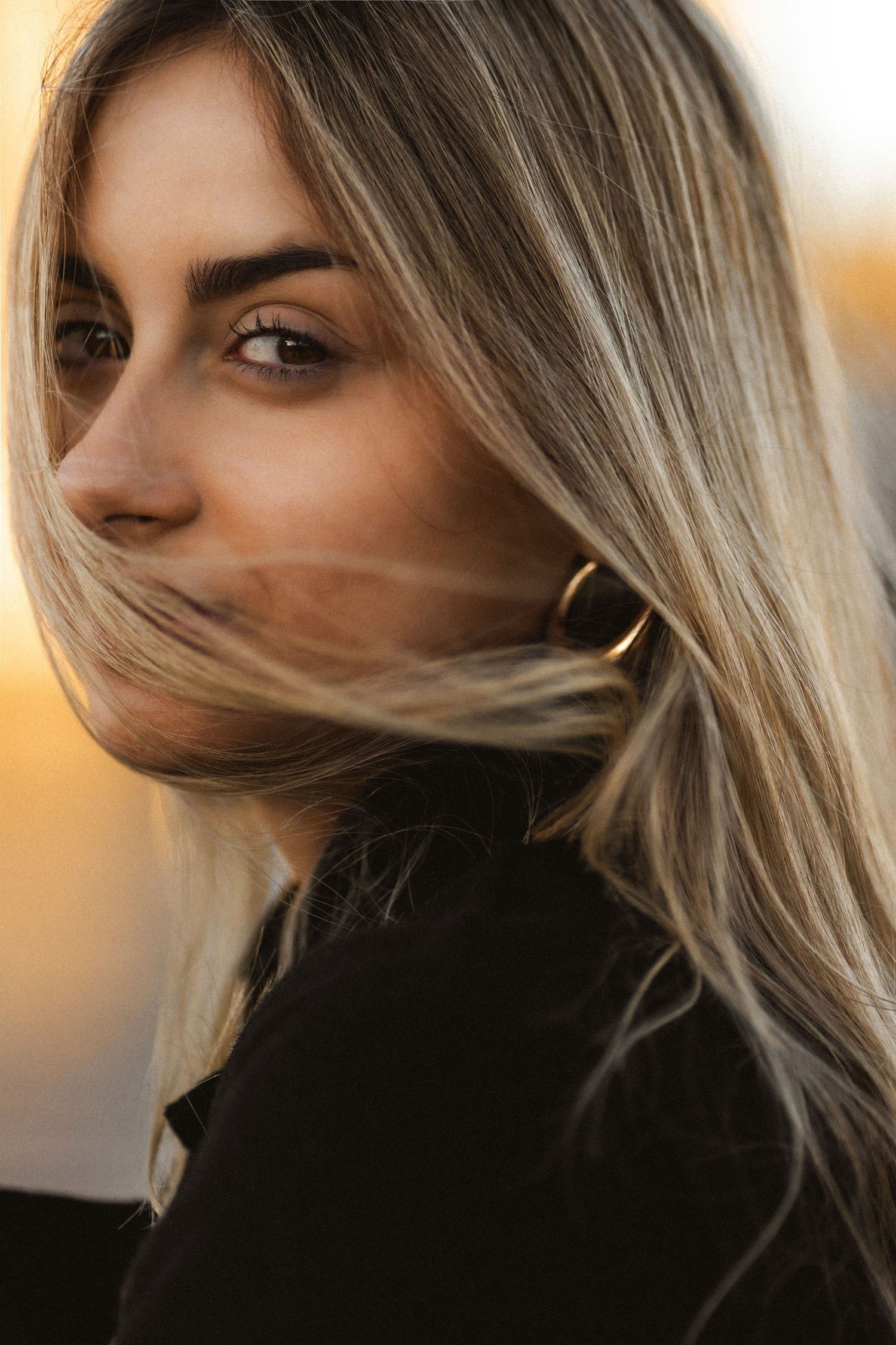 Close-up portrait of someone with blonde hair wearing a black top in warm lighting against a blurred background.