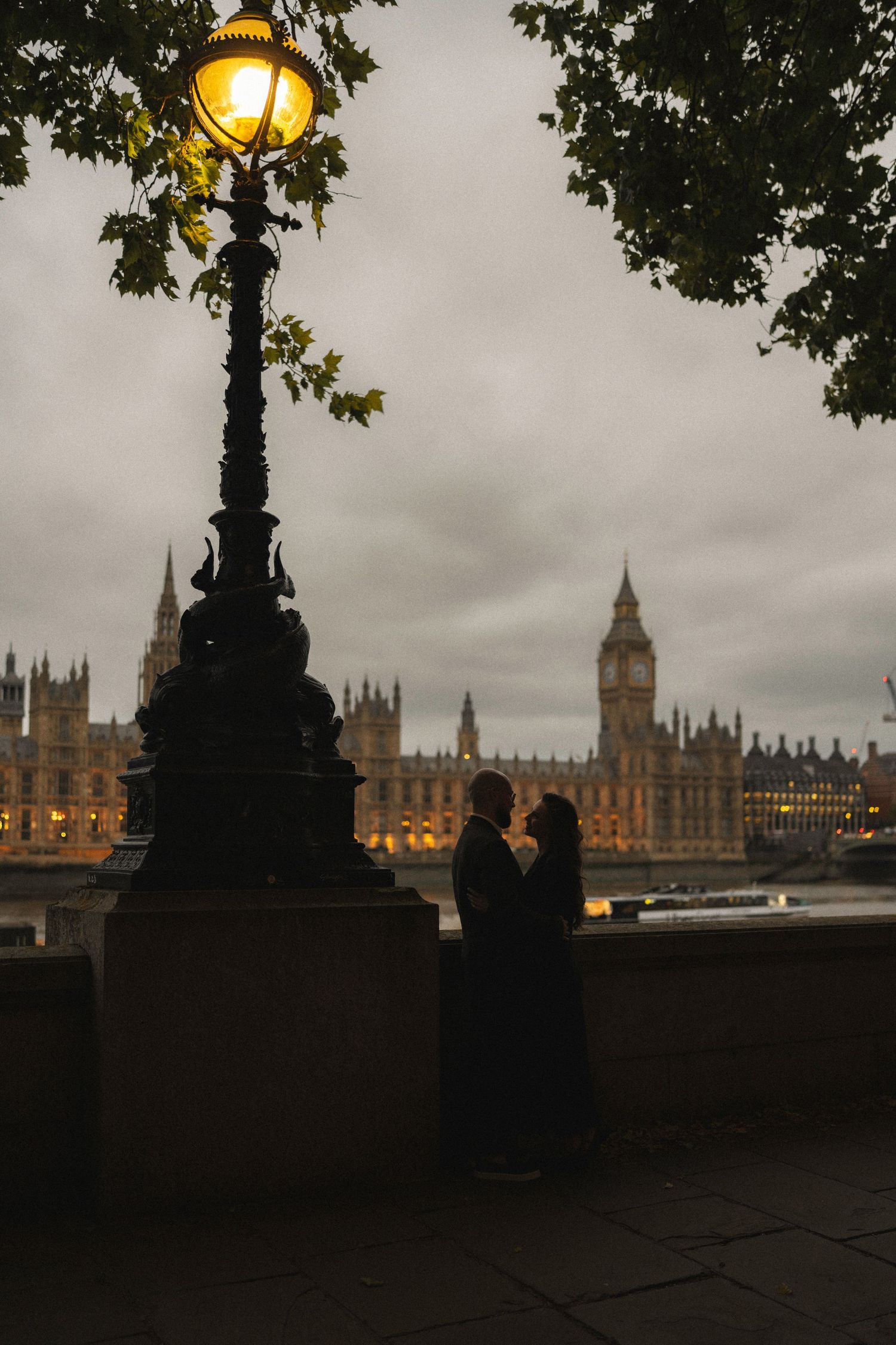Couple silhouetted by glowing lamppost on Thames embankment with Big Ben and Houses of Parliament at dusk.