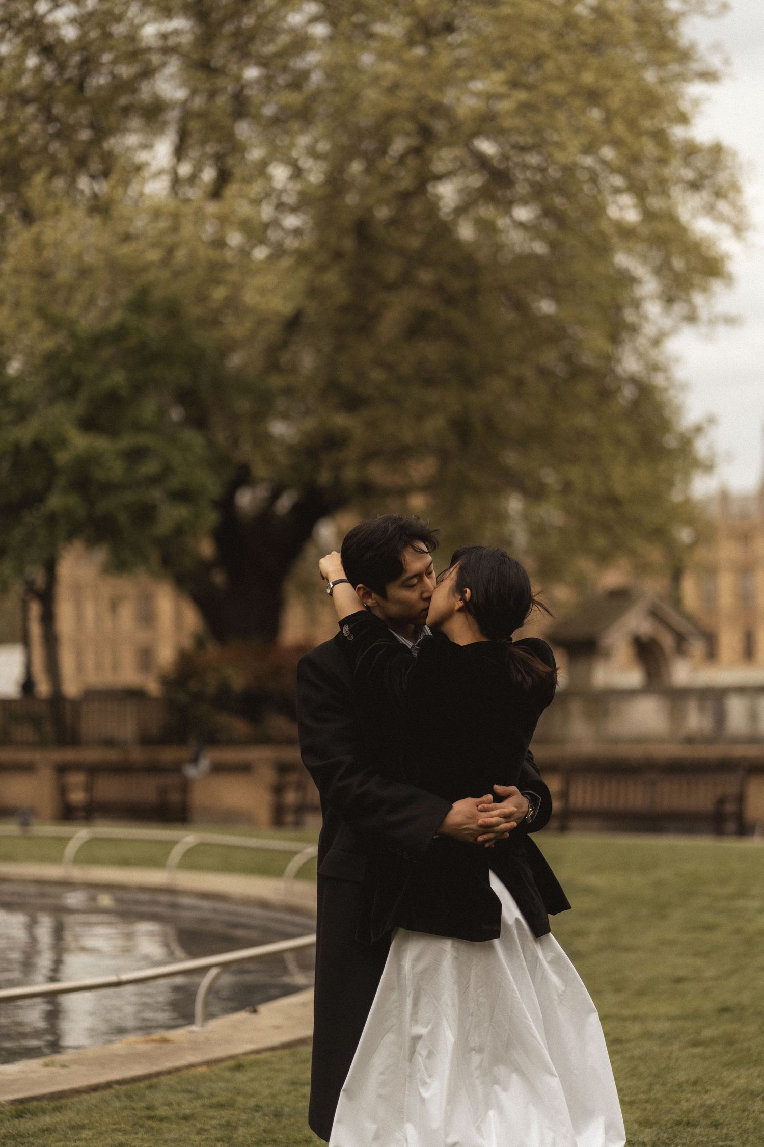 A romantic couple in formal wedding attire embraces on a grassy lawn near a reflecting pool on an overcast day.