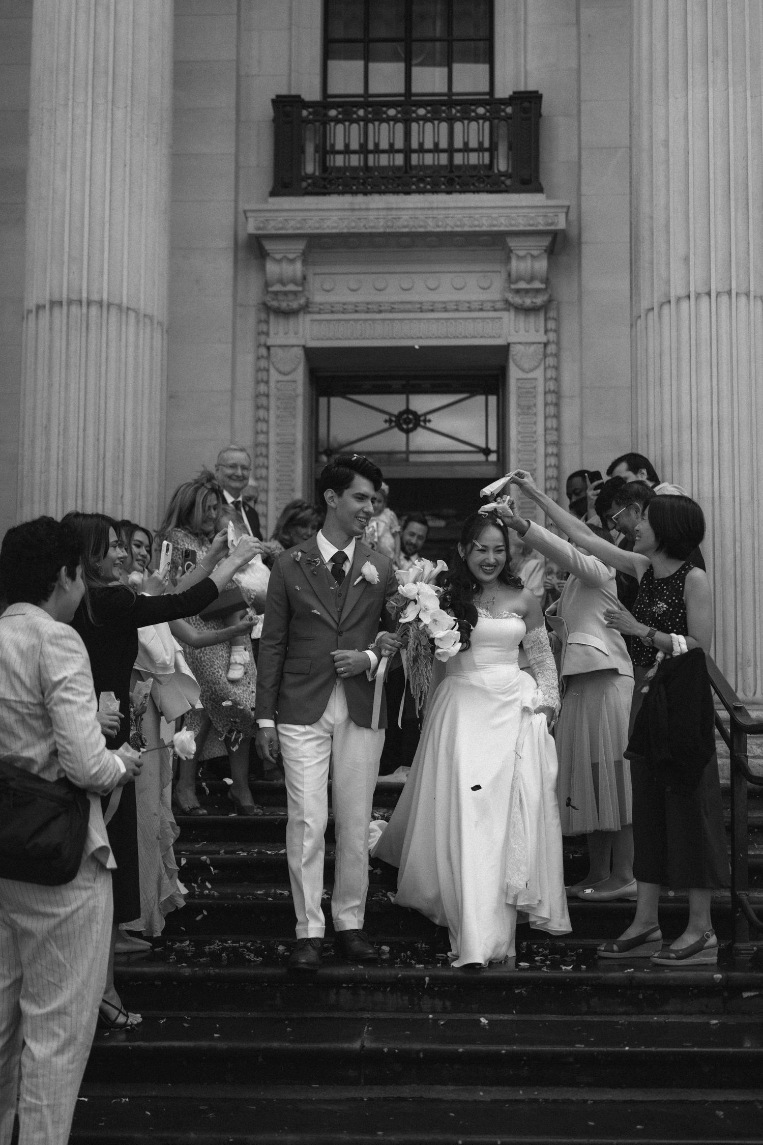 Wedding party celebrates on the steps of a columned building with guests throwing rice and confetti in black and white.