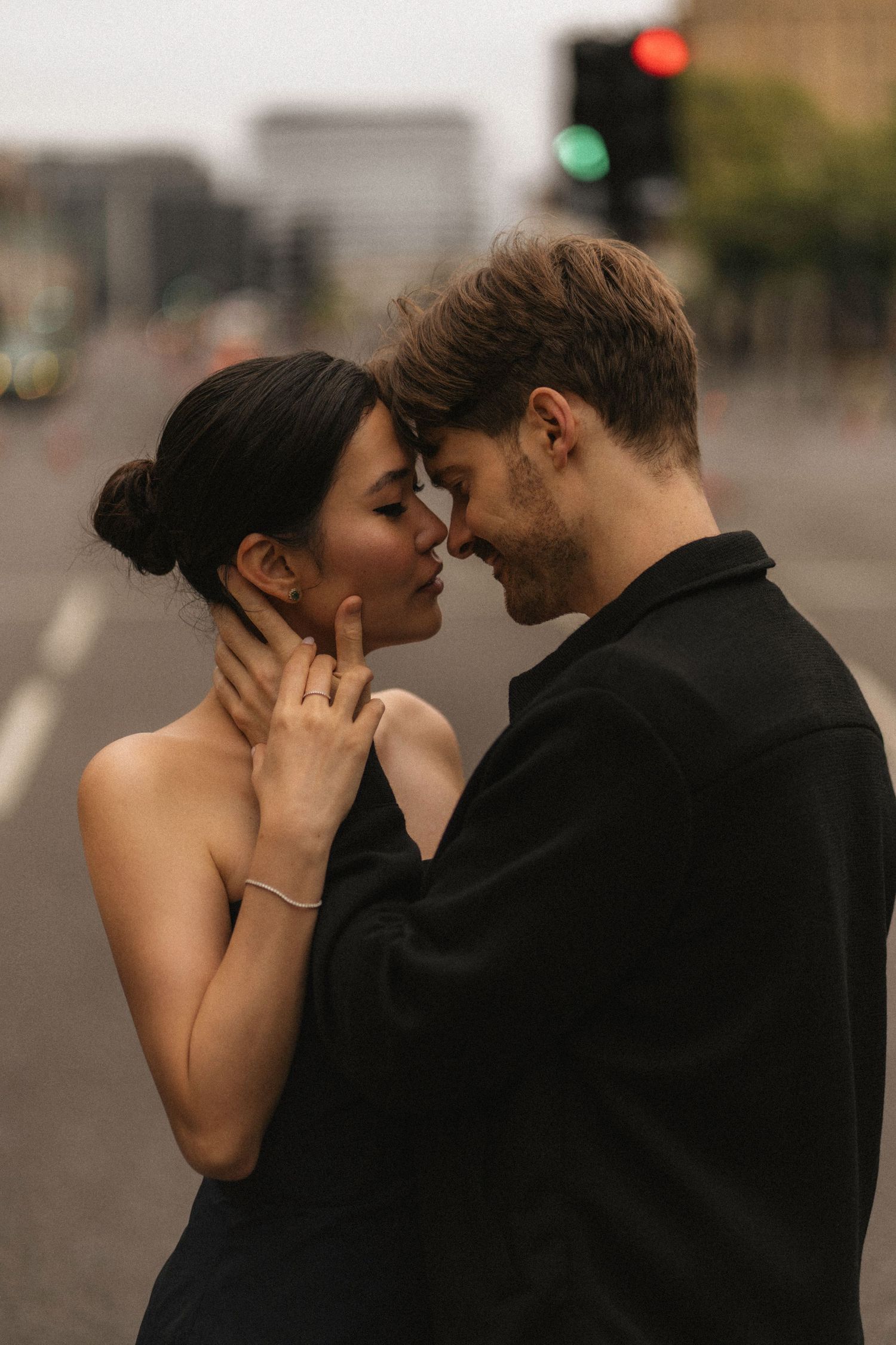 A romantic couple in black attire share an intimate kiss on a city street at dusk.
