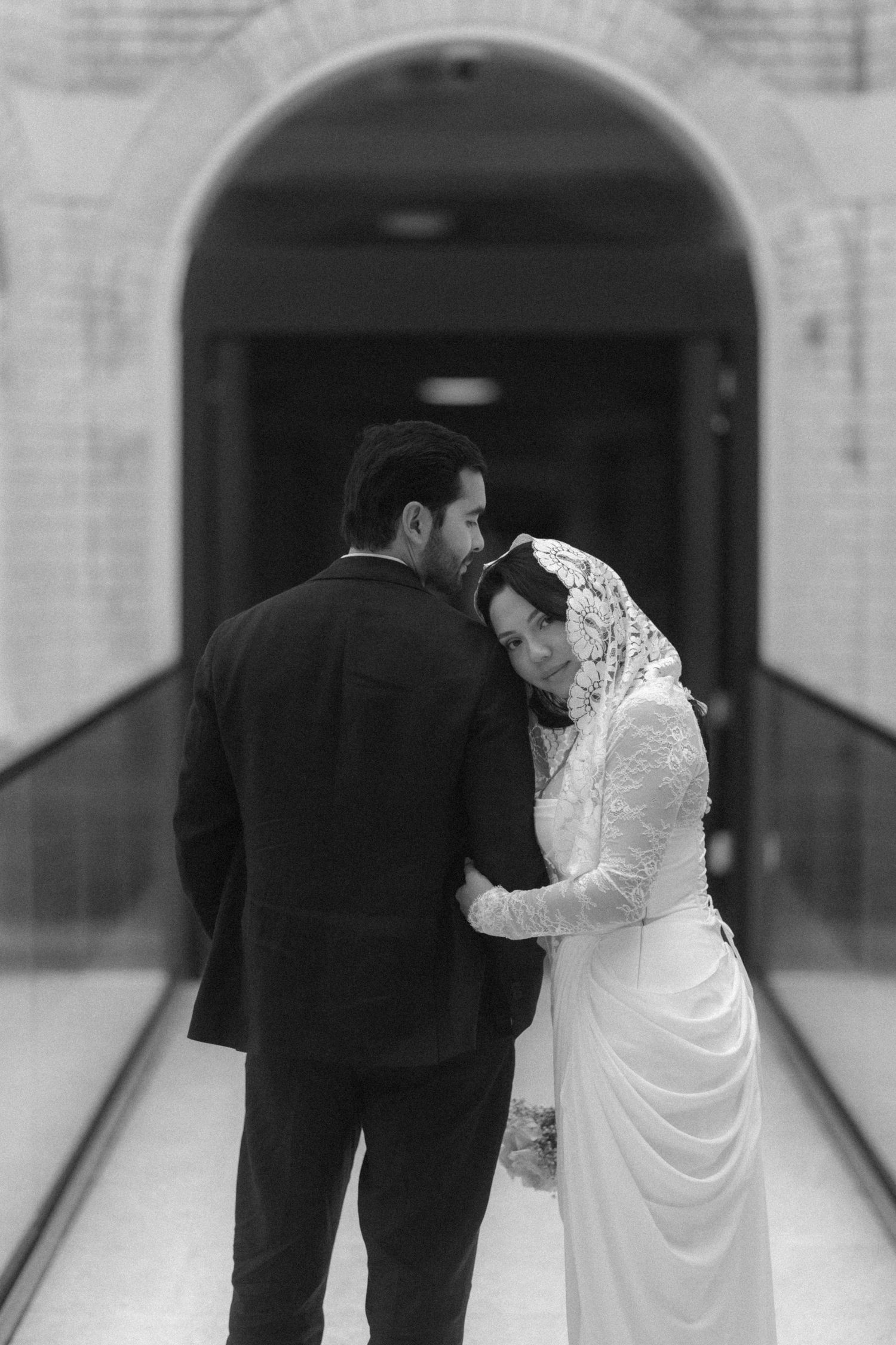 Black and white photo of a Muslim bride and groom embracing tenderly in an elegant arched hallway on their wedding day.