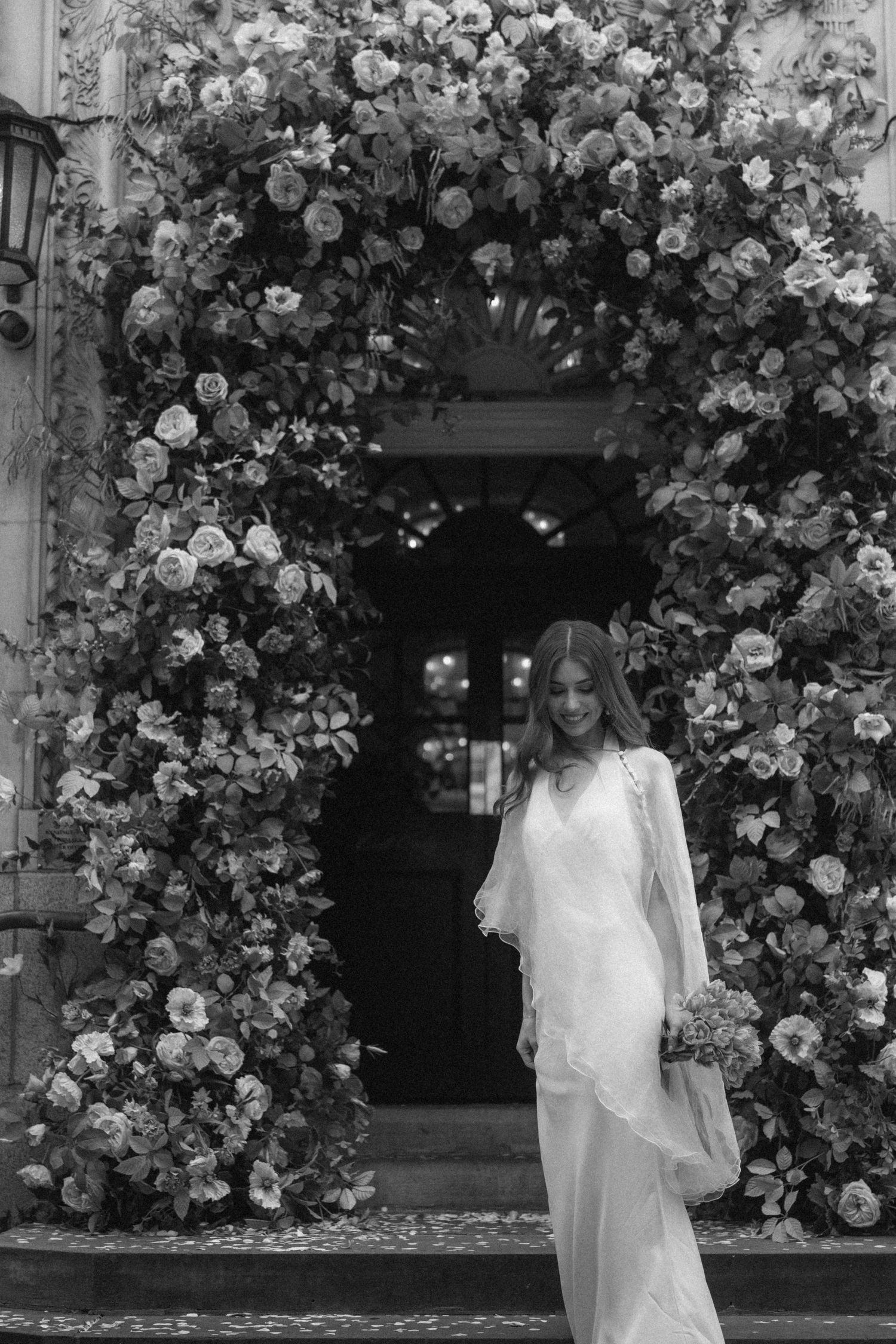 Bride in white gown stands under floral arch doorway in black and white wedding photo.