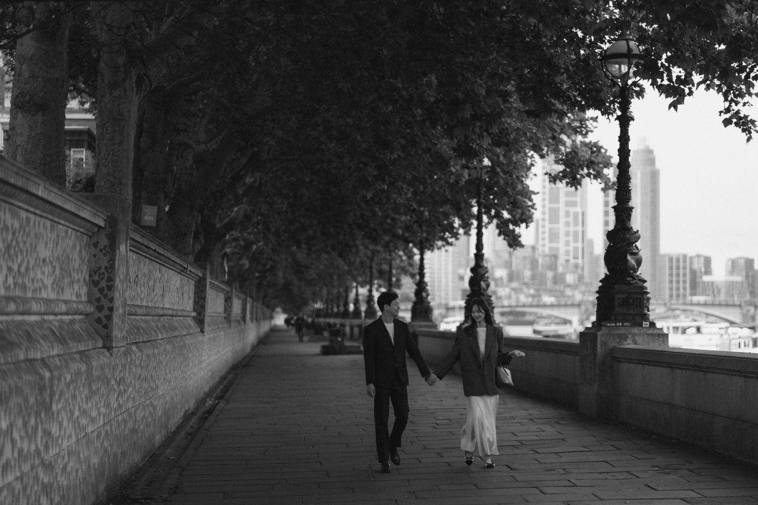 Black and white photo of people walking along Thames embankment in London with city skyline in background.