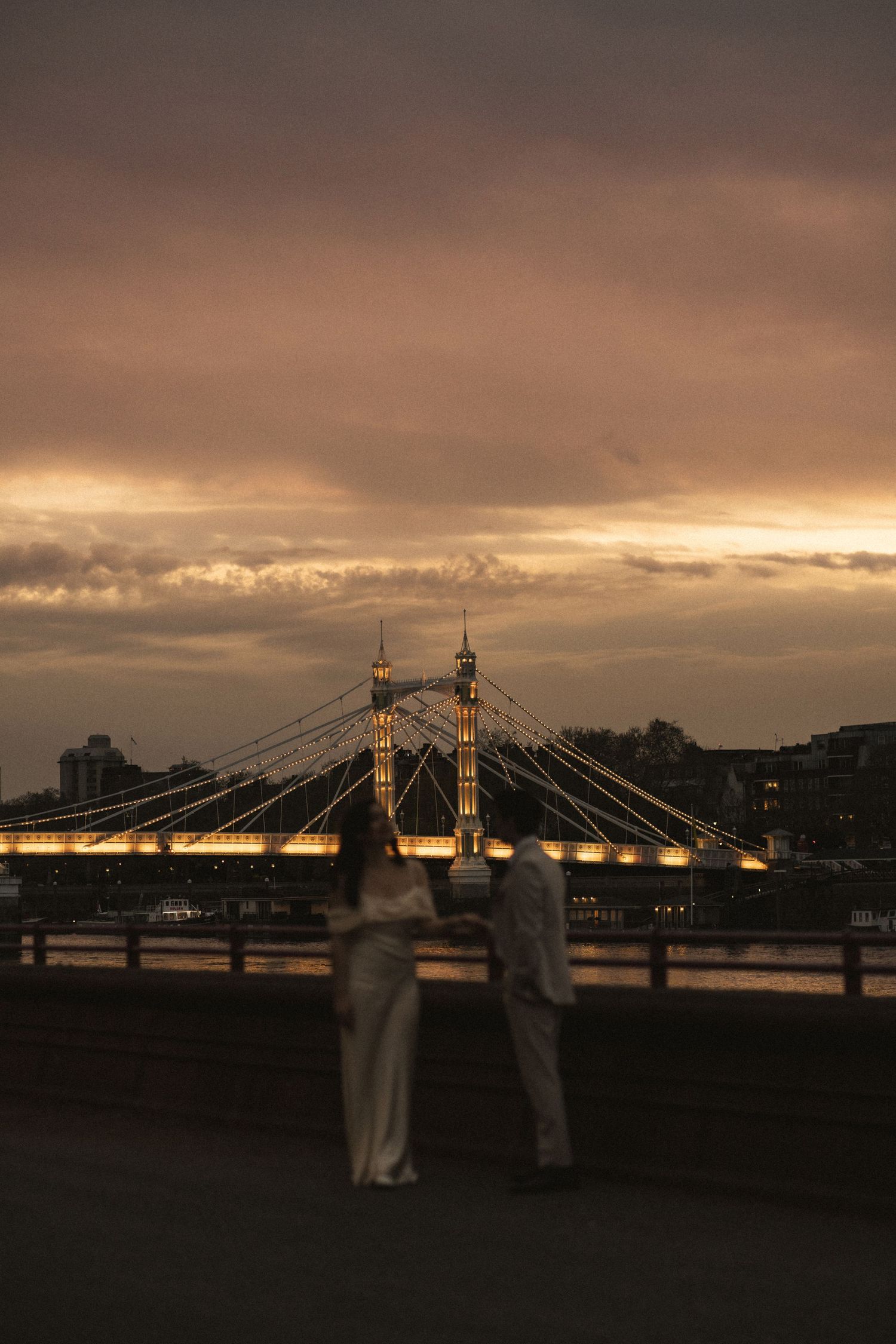 Two silhouettes admire Albert Bridge in London at sunset with dramatic orange clouds illuminating the iconic structure.