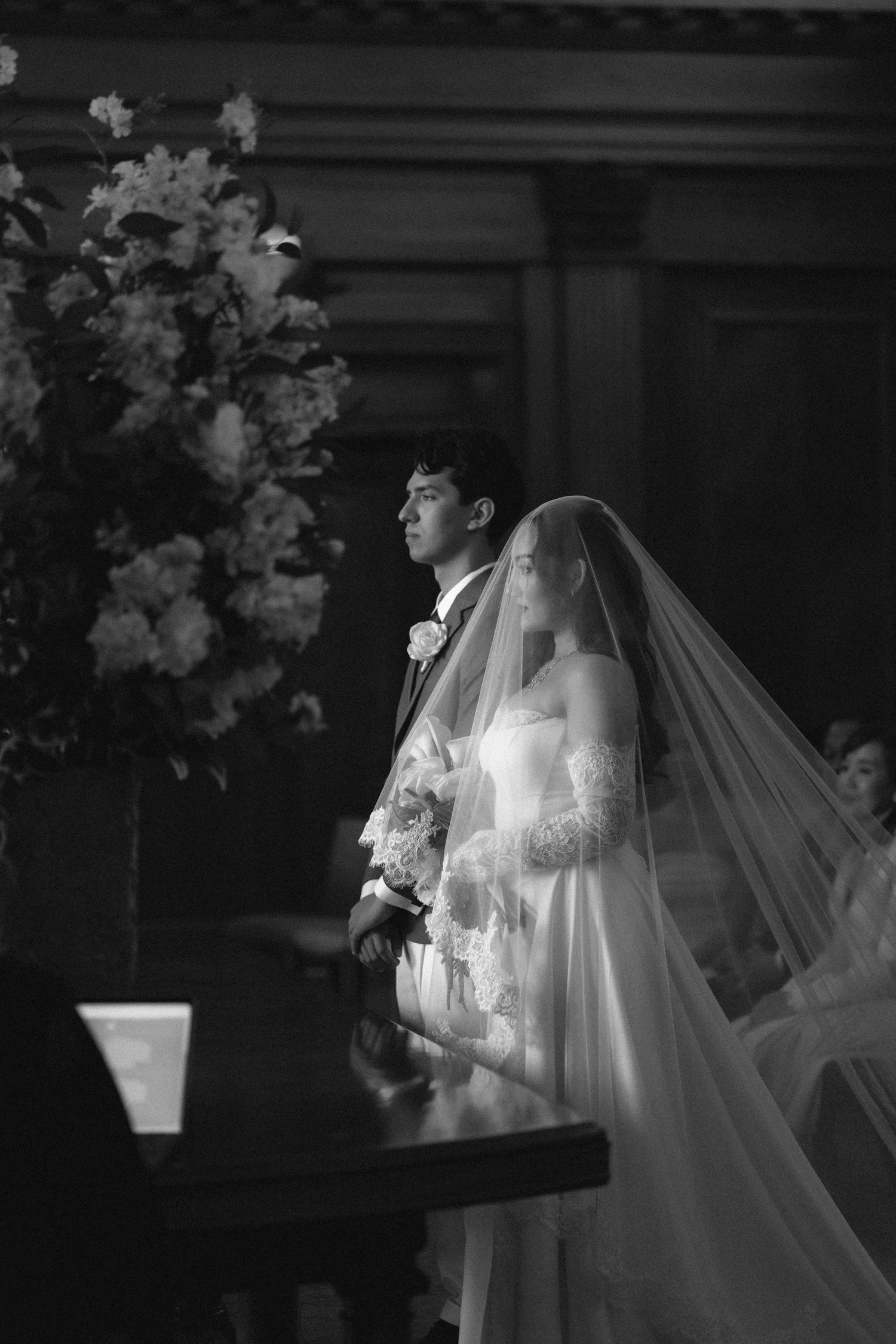 A bride in a flowing white wedding gown and long veil holds a bouquet in a dramatic black and white photograph.