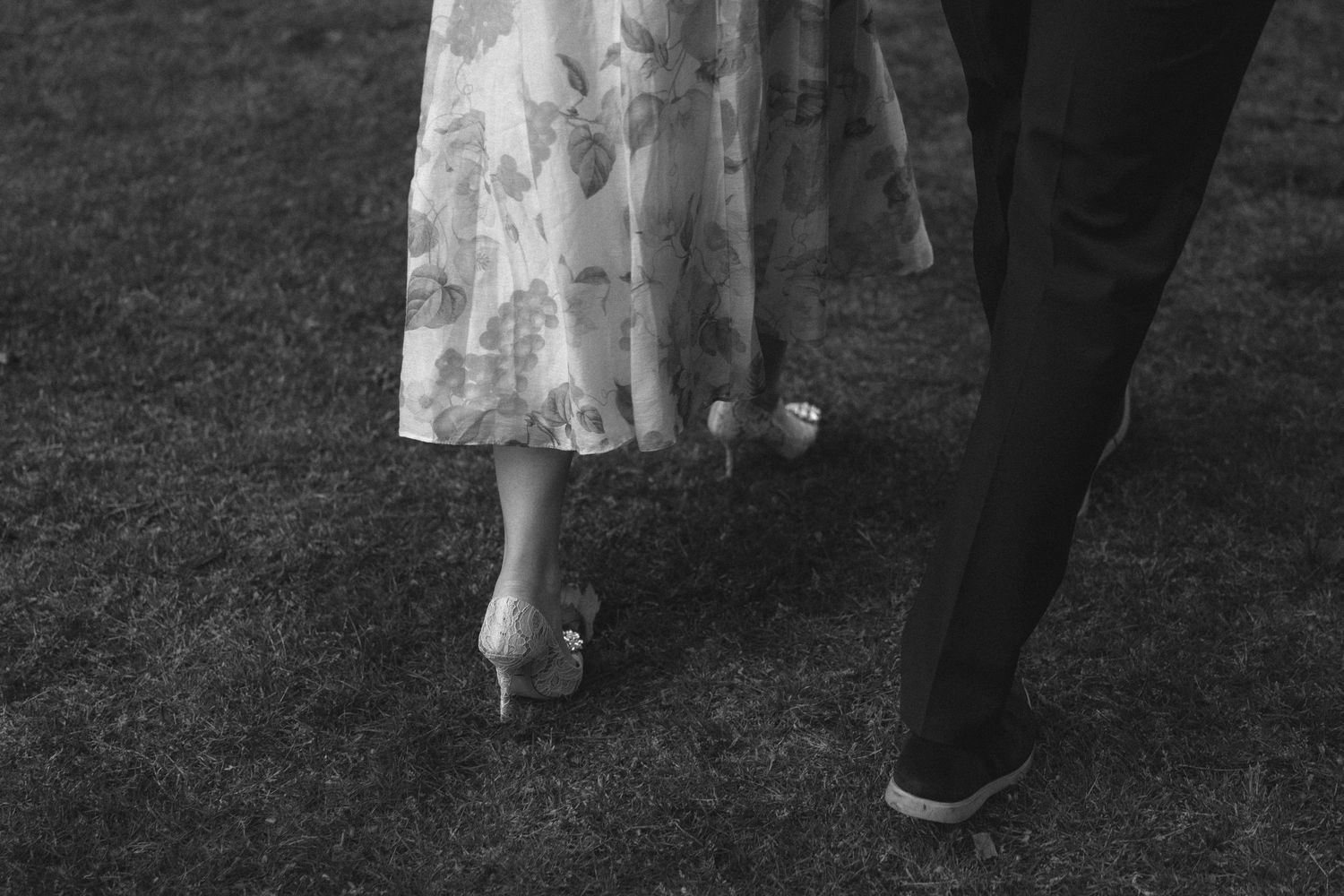 A person's legs in black pants and sneakers walking on grass next to a white dress hem.