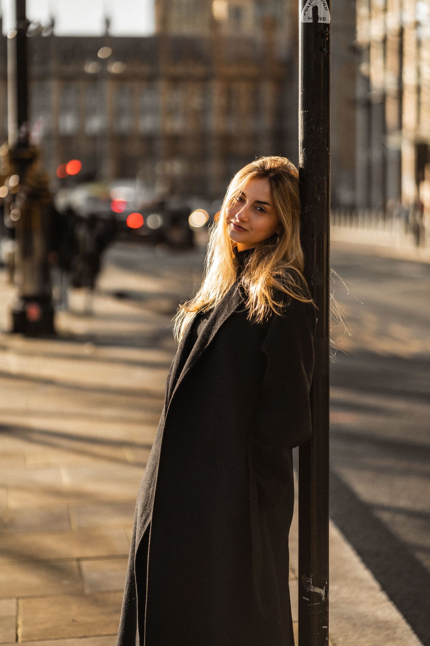 A person in a black coat poses against a city backdrop during golden hour lighting.
