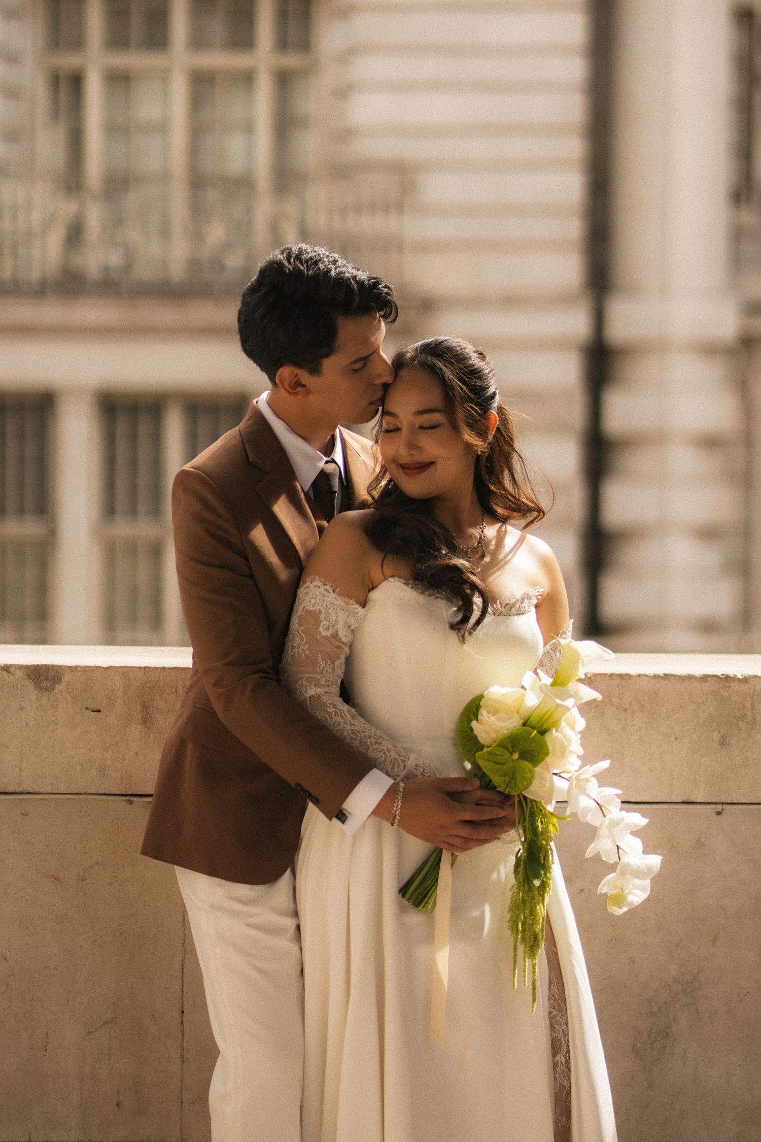 A romantic wedding couple embraces on a balcony as the groom kisses the bride holding a bouquet of white calla lilies.