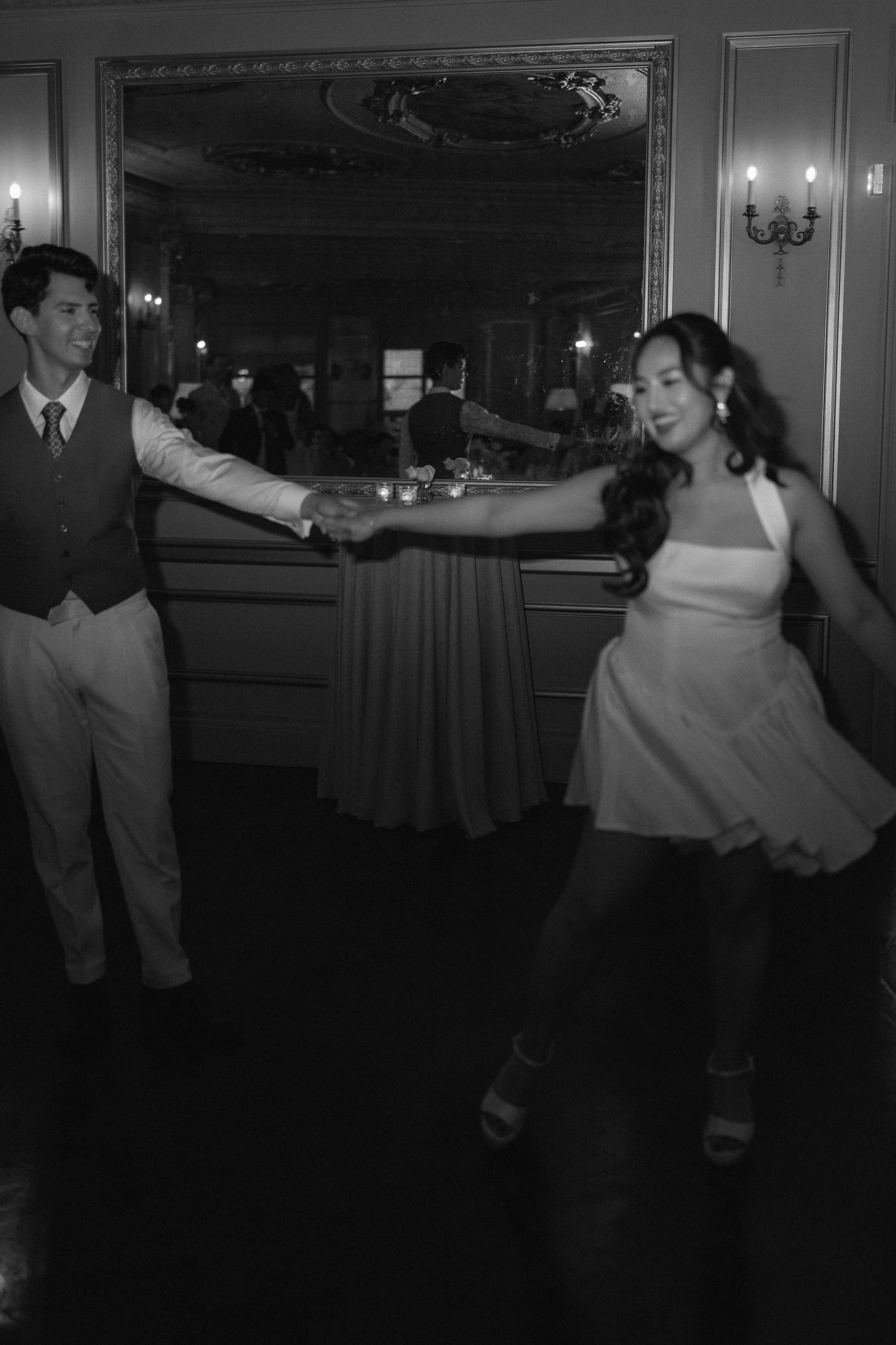 Couple dancing elegantly in black and white photo at a formal event, man in vest holds woman in white dress hand.