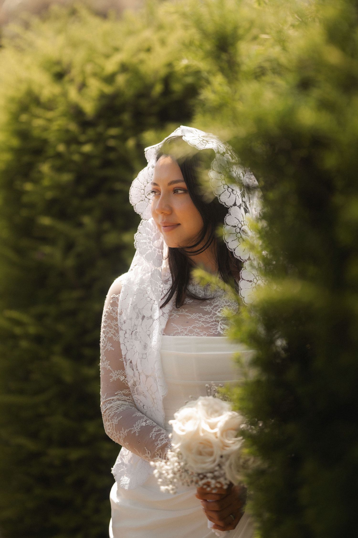 Bride in lace veil and white gown holding bouquet of white roses surrounded by lush green hedges.