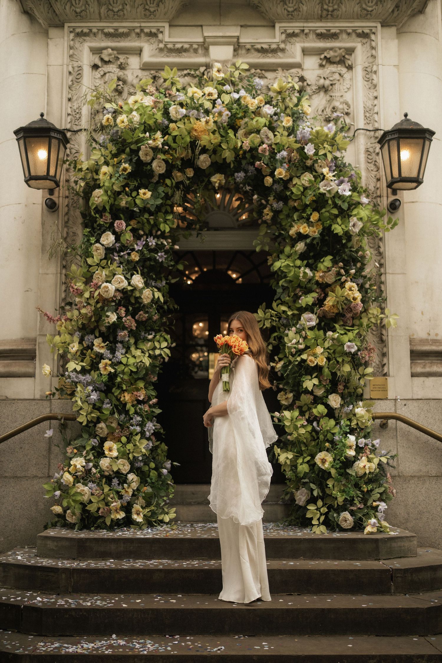 Bride in white gown standing beneath lush floral arch at ornate building entrance with lanterns.