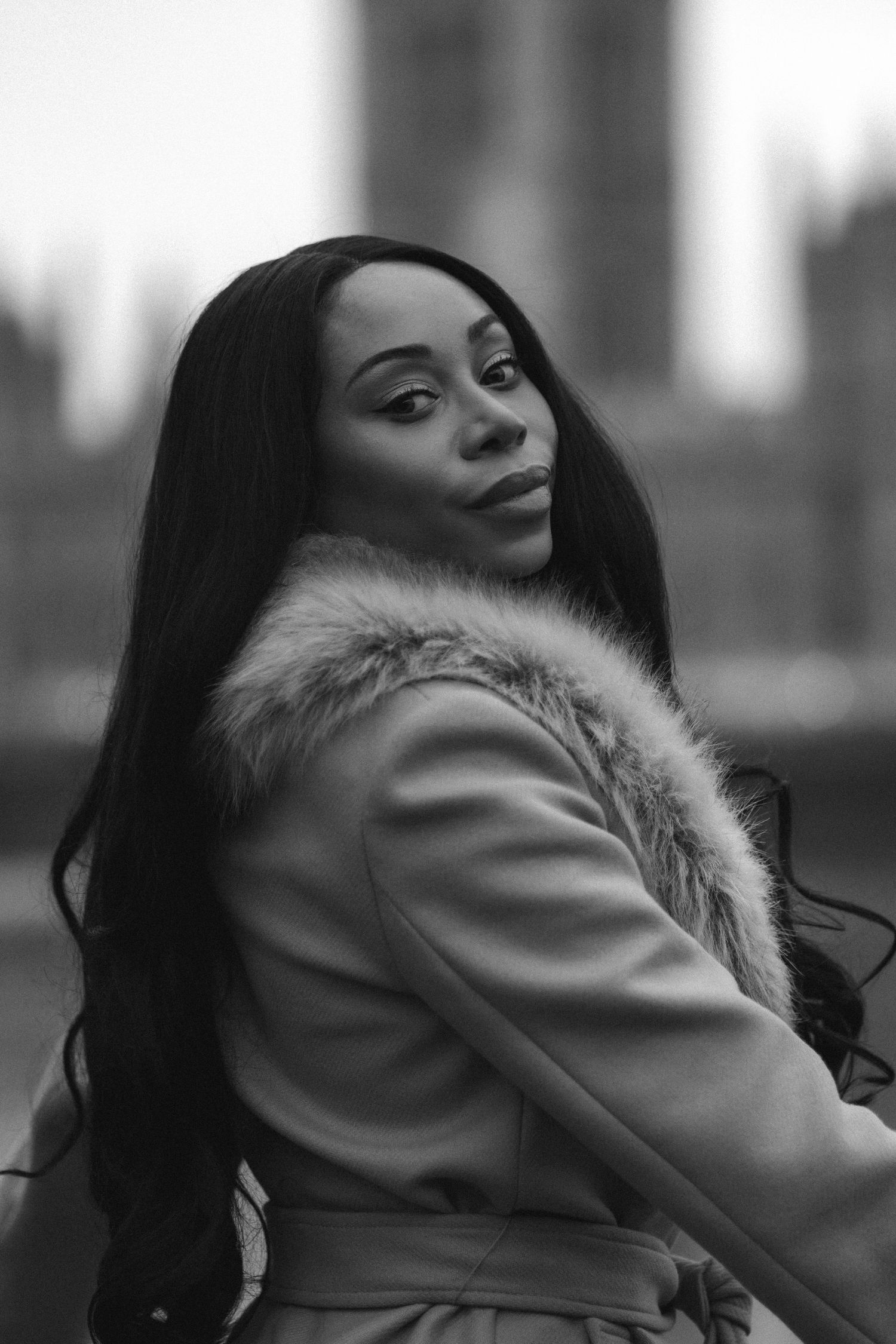 A woman in a fur-trimmed coat poses dramatically against a city backdrop in black and white photography.