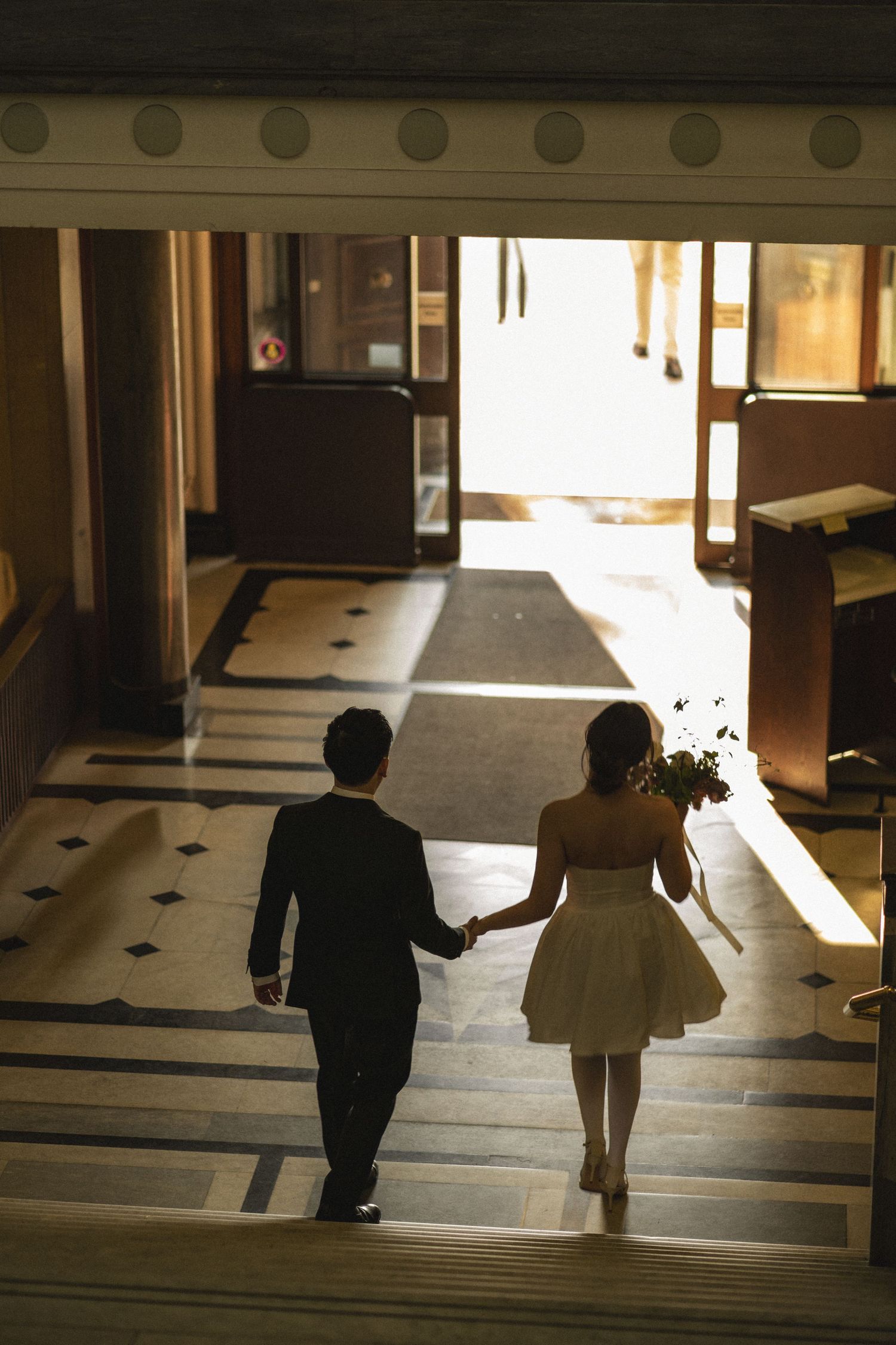 Bride and groom holding hands walking toward sunlit entrance in elegant venue with marble floors.