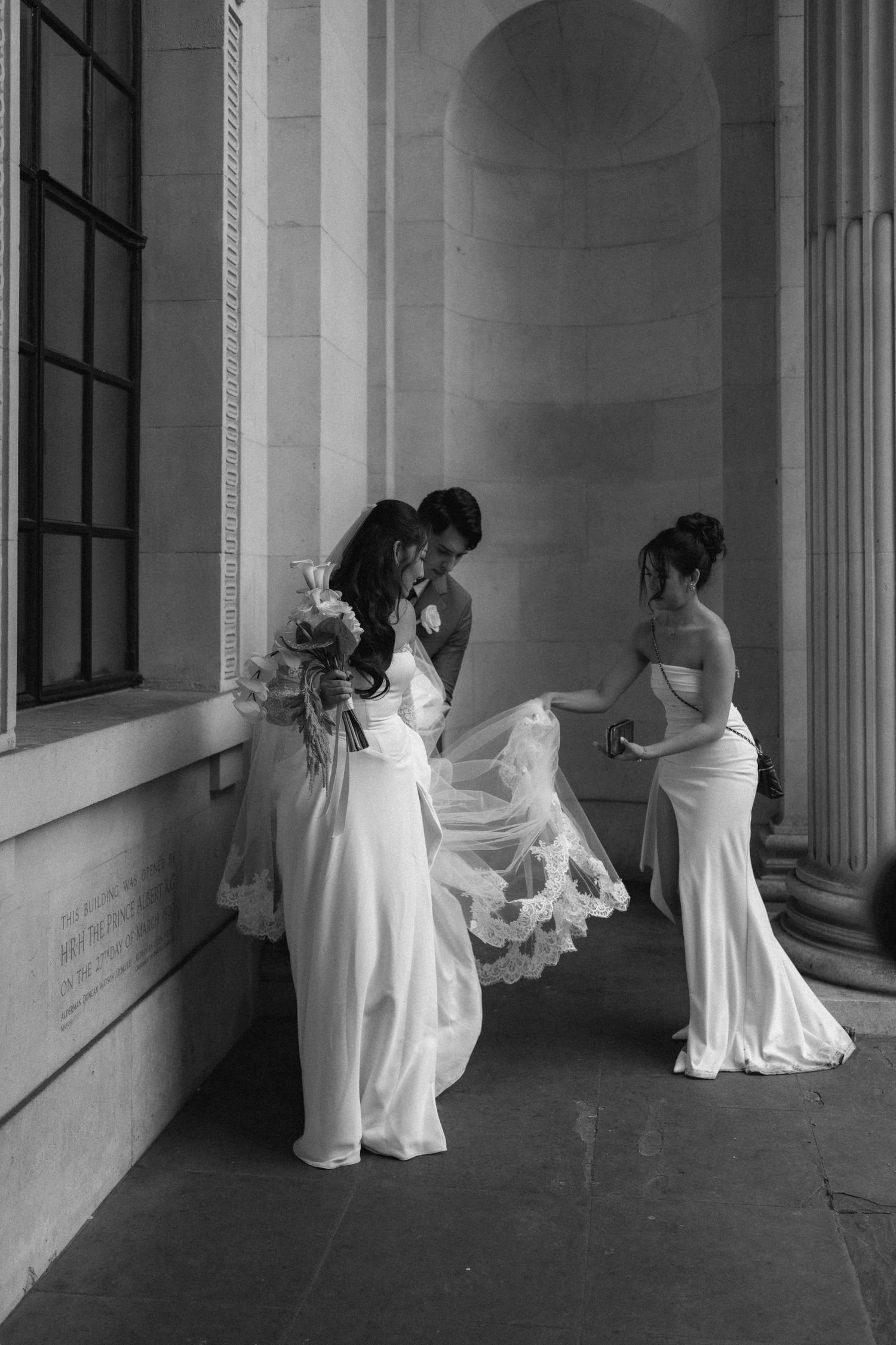 Black and white photo of a bride and groom sharing a kiss while a bridesmaid adjusts the veil outdoors.