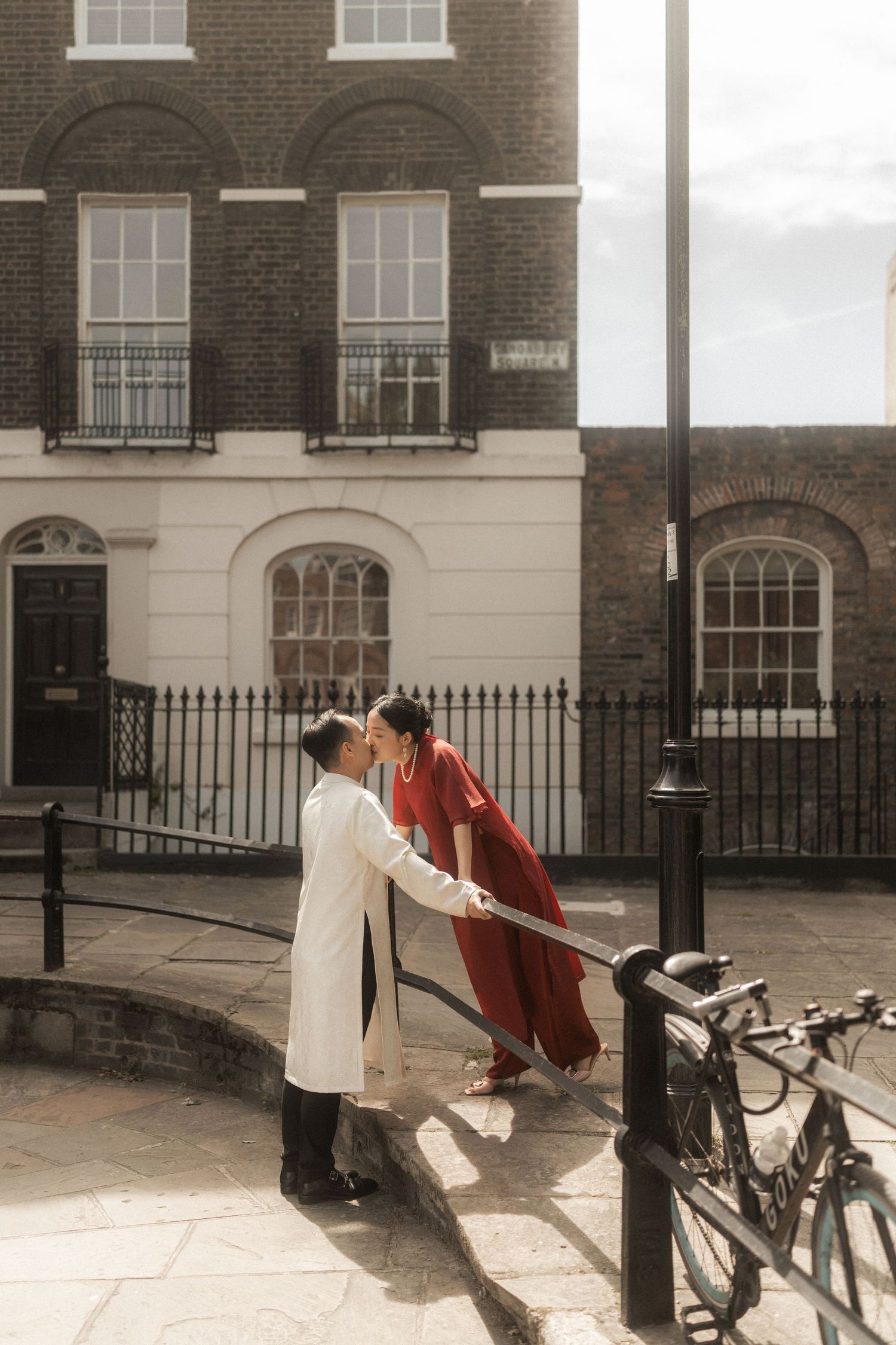 Couple in red and white traditional attire holding hands by iron railings on a sunlit London street.
