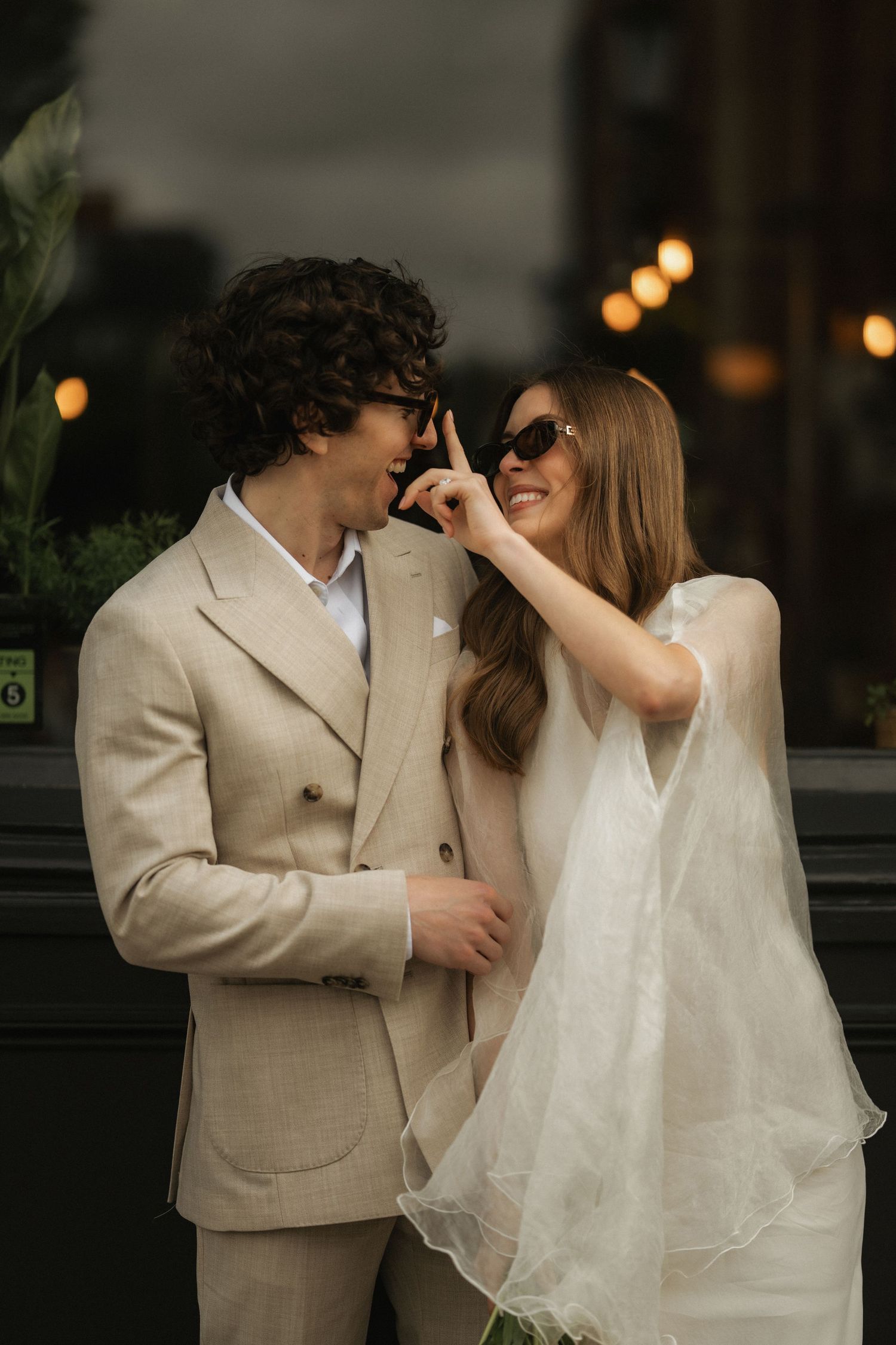 A smiling bride in a white dress playfully adjusts sunglasses on a groom in a beige suit outdoors.