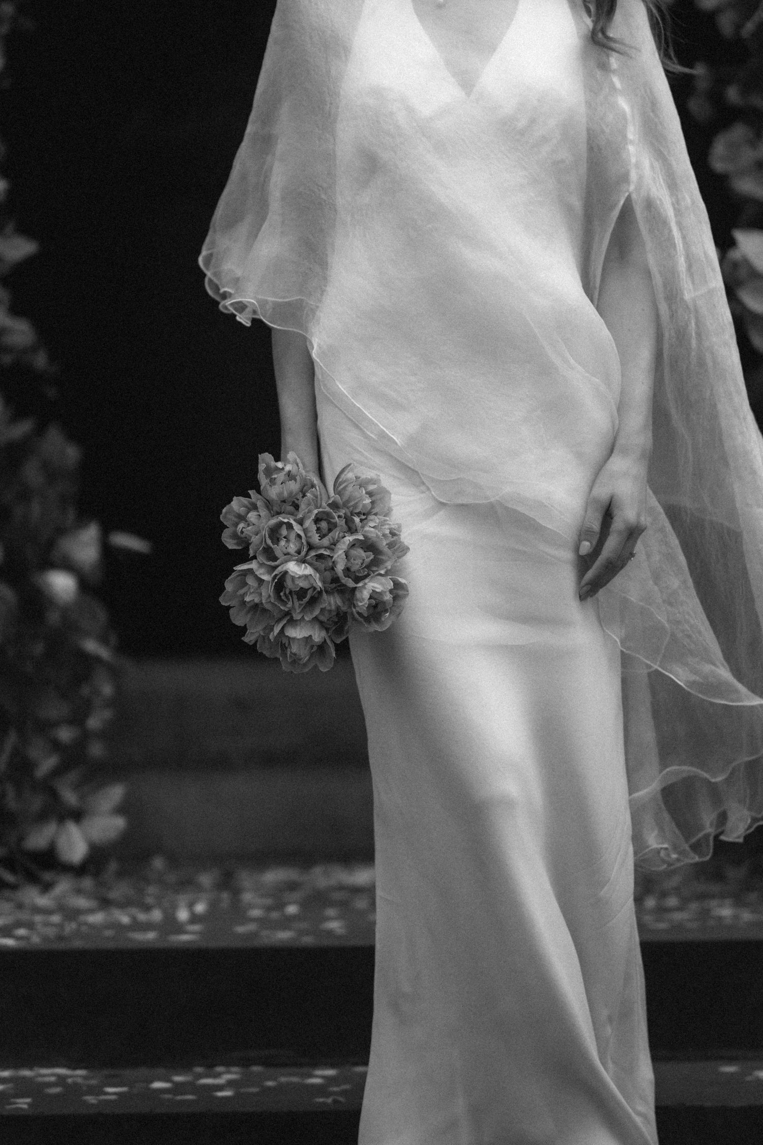 Black and white photo of a bride in a silk gown and veil holding a floral bouquet.