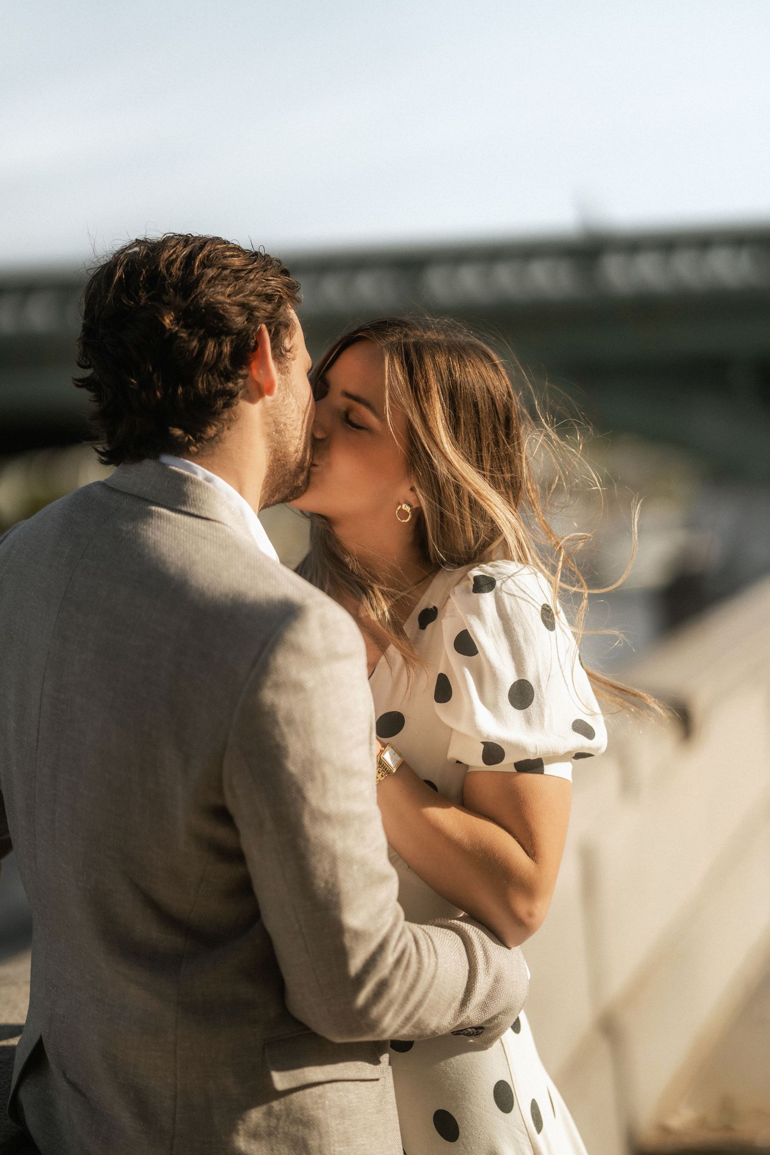 Couple sharing a romantic kiss outdoors, woman in polka dot dress embraced by man in gray suit.