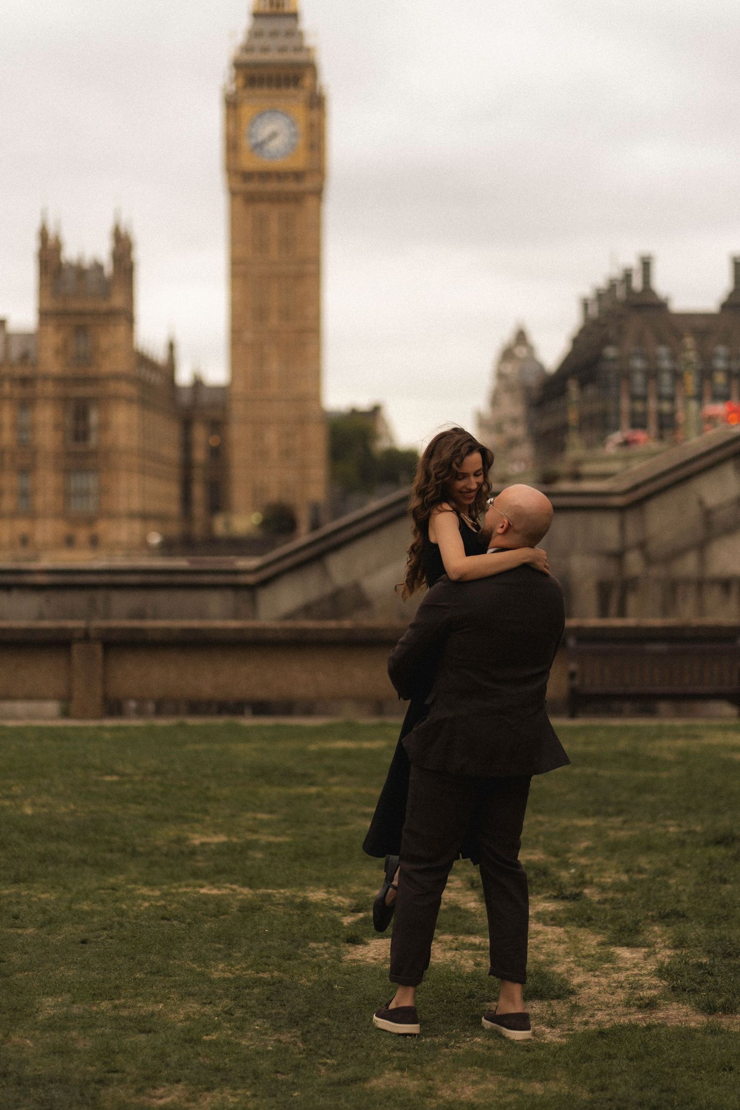 A couple embraces in front of Big Ben in London, dressed in black on a cloudy day.