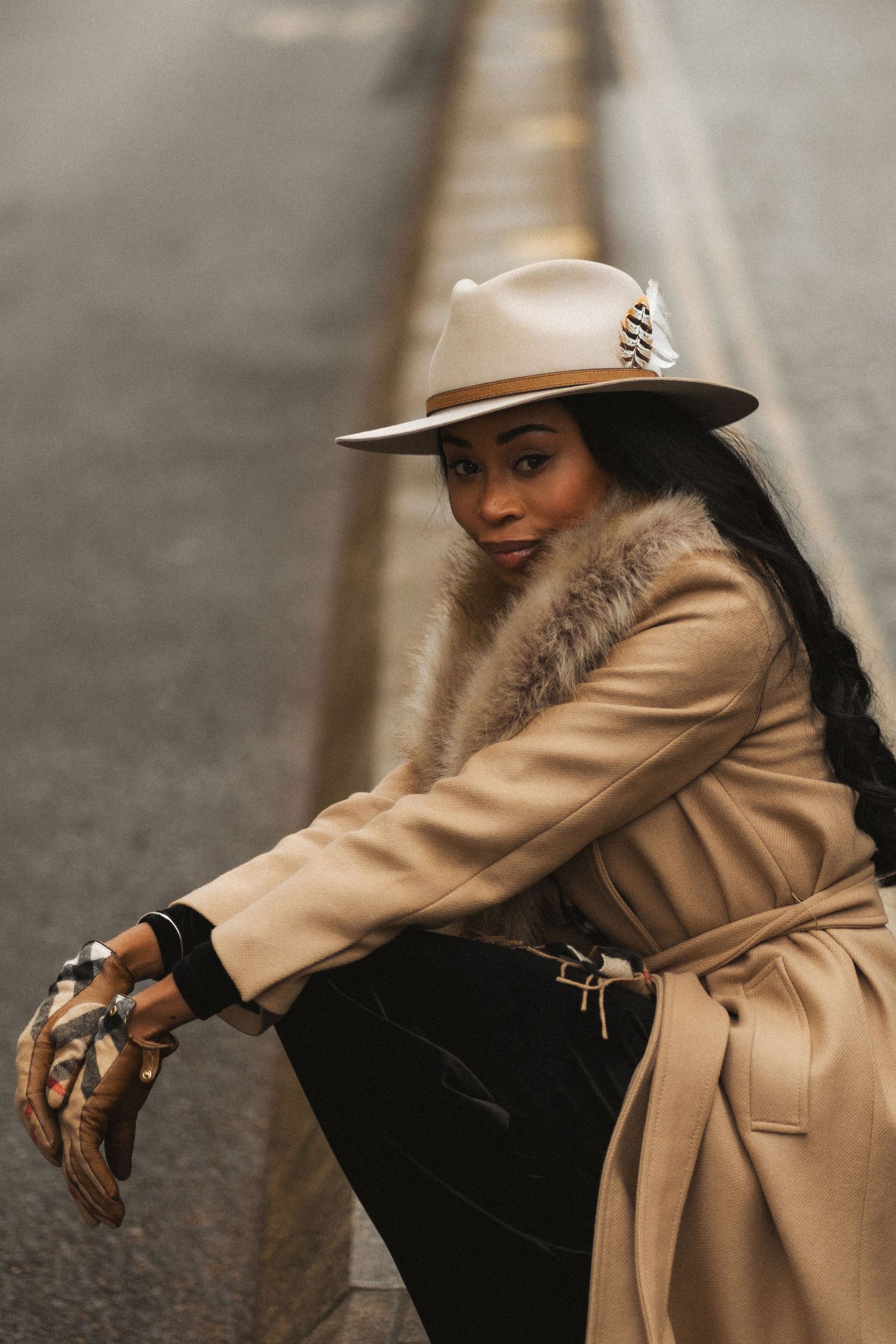 A person in a beige coat and white hat crouches down in a dynamic fashion-forward pose on a street.