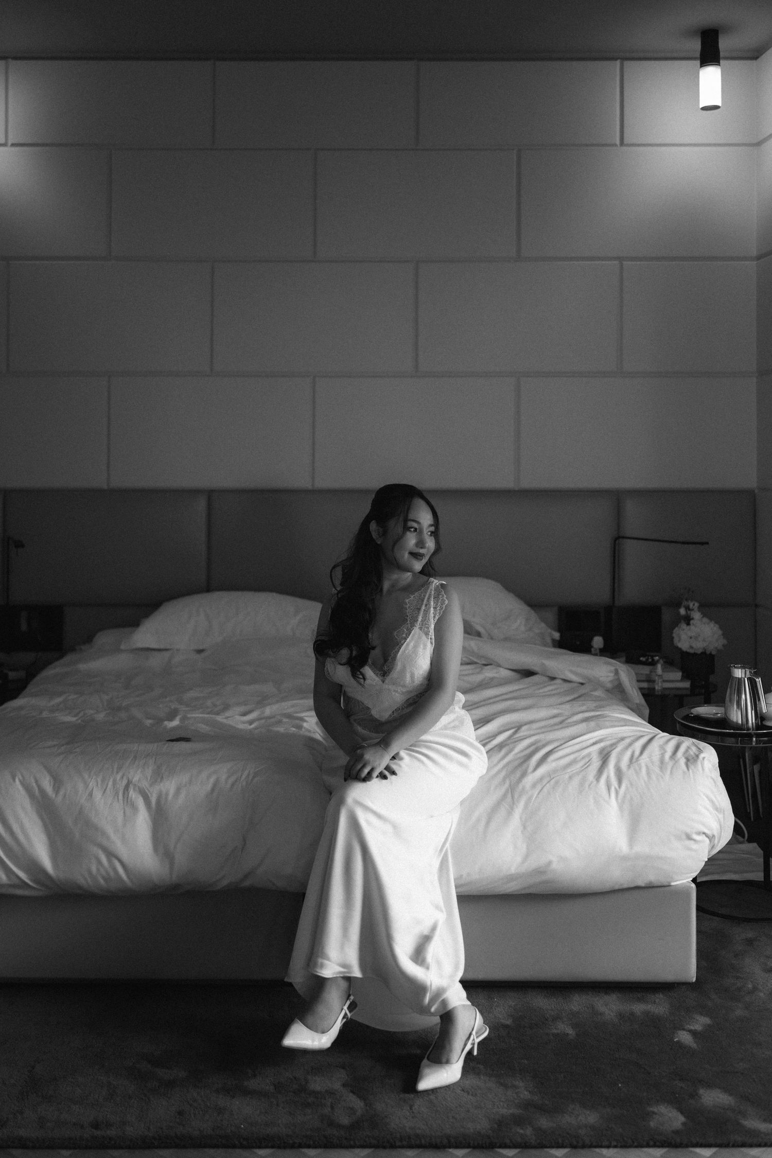 Black and white photo of a woman in a white dress sitting on a hotel bed, gazing thoughtfully to the side.